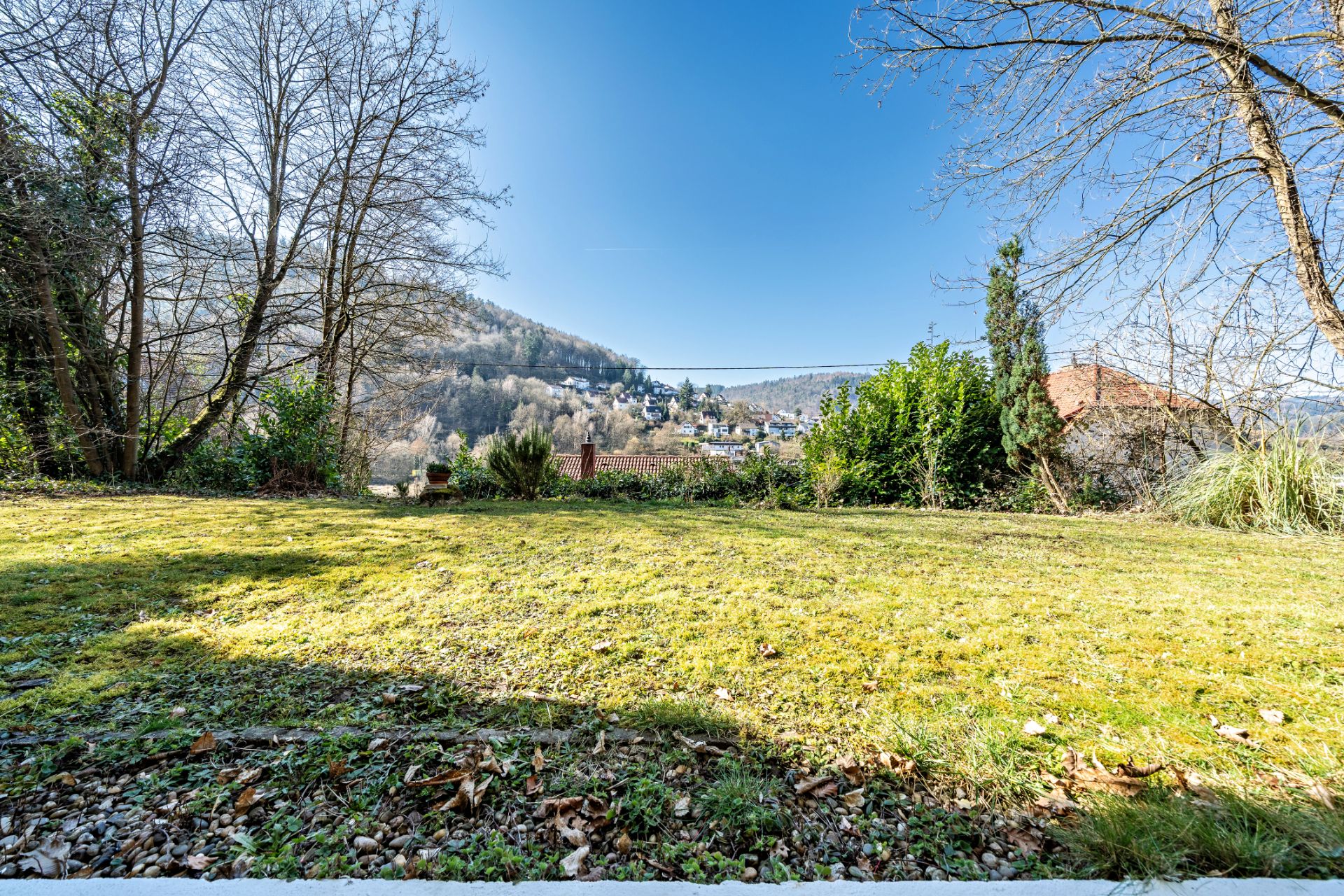 Garten mit Rasen, Bäumen und Blick auf umliegende Hügel und Häuser unter blauem Himmel.