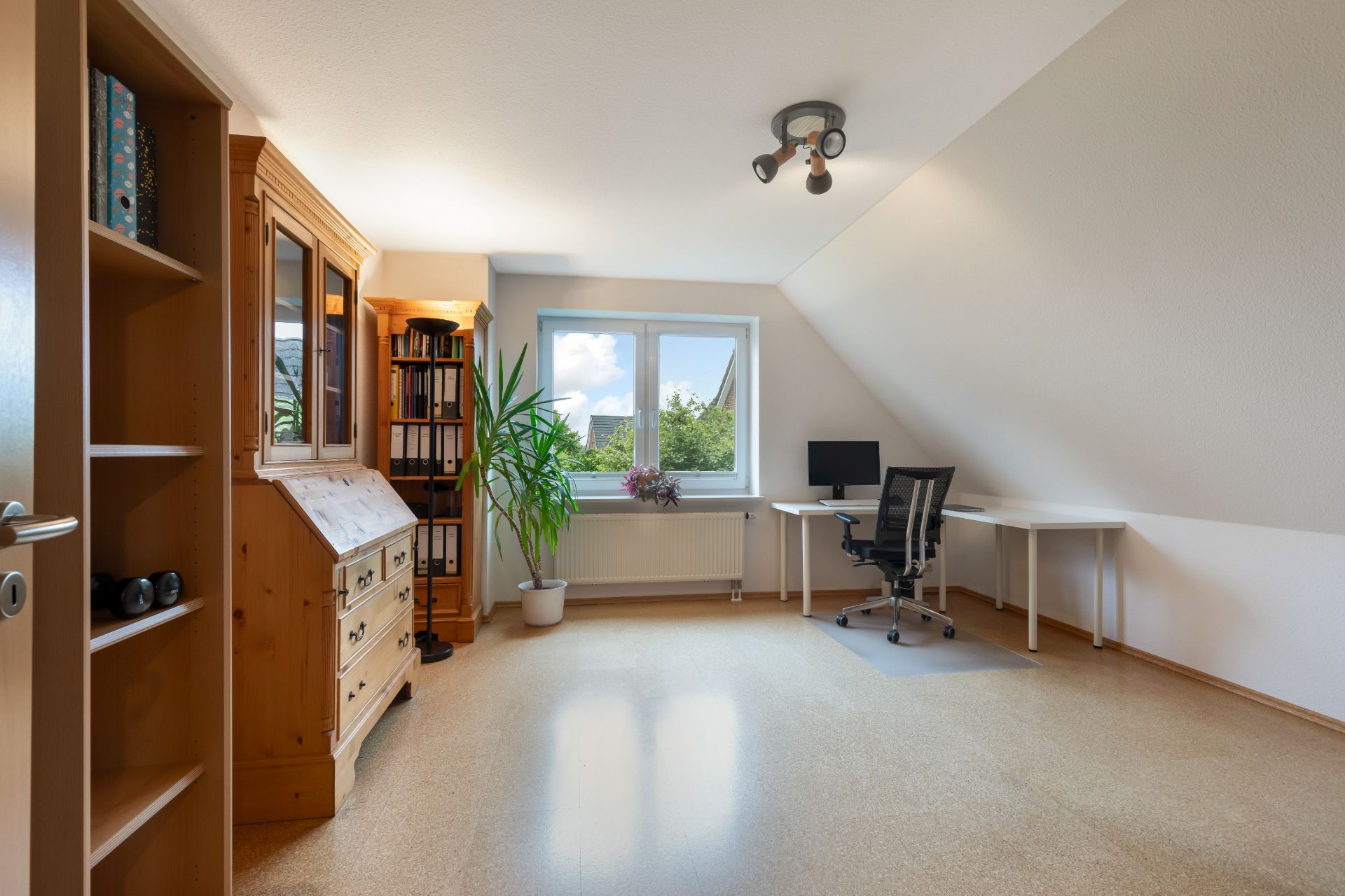 Helles Arbeitszimmer mit Schreibtisch, Stuhl, Regal, Schrank, Pflanze und Fenster.