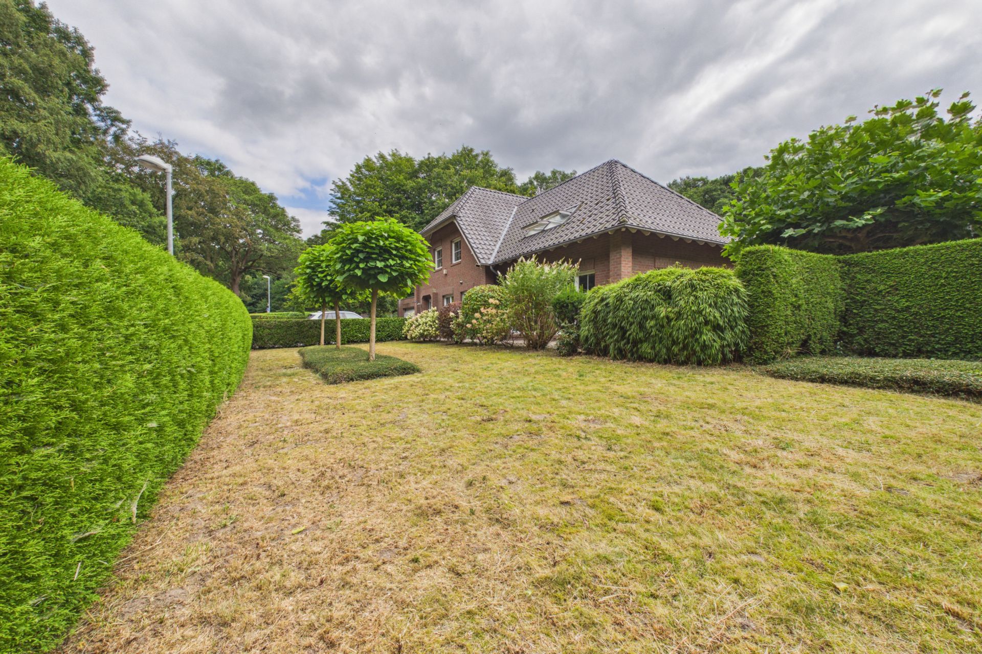 Backsteinwohnhaus mit gepflegtem Garten, Hecken und kleinen Bäumen bei bewölktem Himmel.