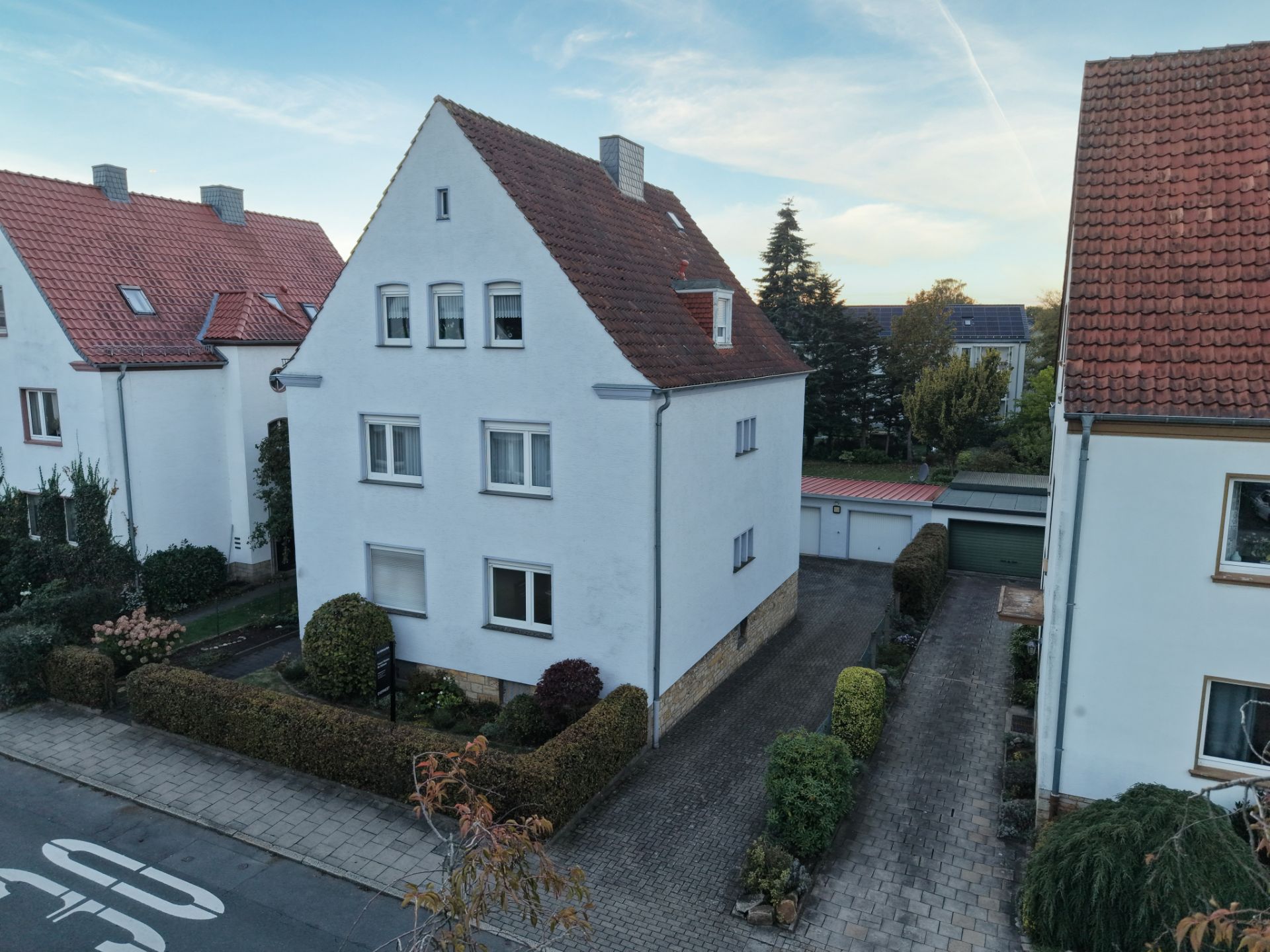 Weißes Wohnhaus mit rotem Dach, Vorgarten und Zufahrt zu Garagen.
