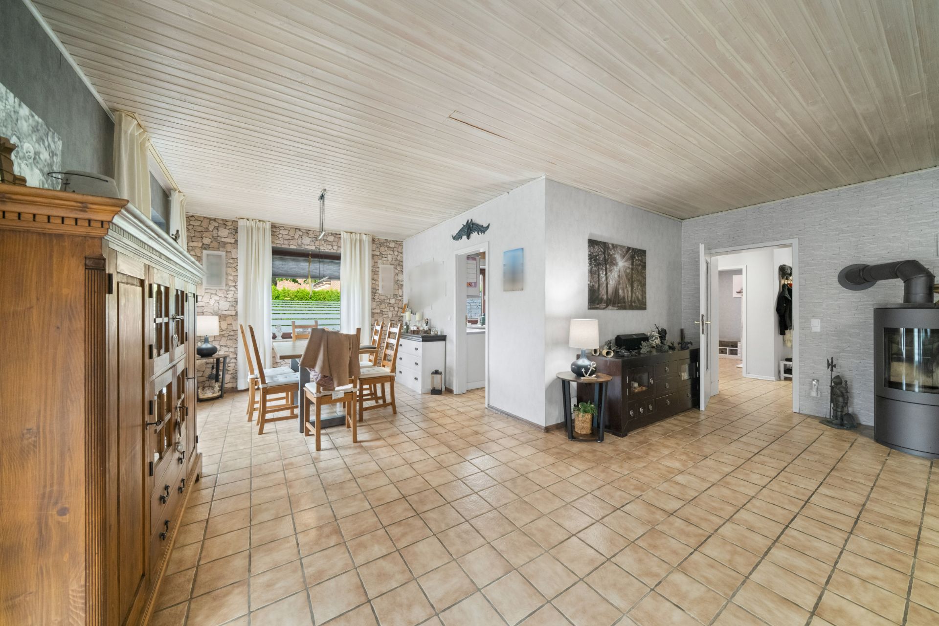 Helles Wohnzimmer mit Esstisch, Holzmöbeln, Fliesenboden und einem Kaminofen.