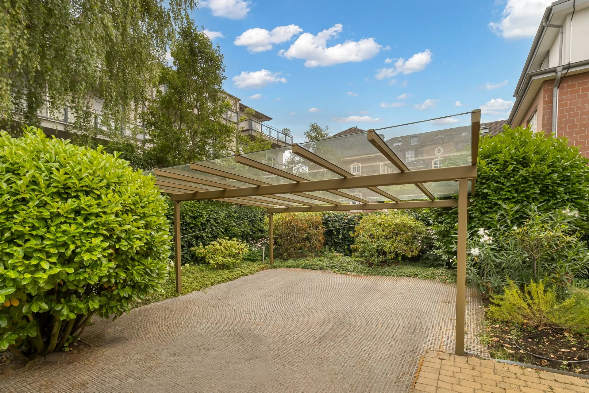Überdachter Carport mit Glasdach und umliegenden Büschen im Gartenbereich eines Wohnhauses.