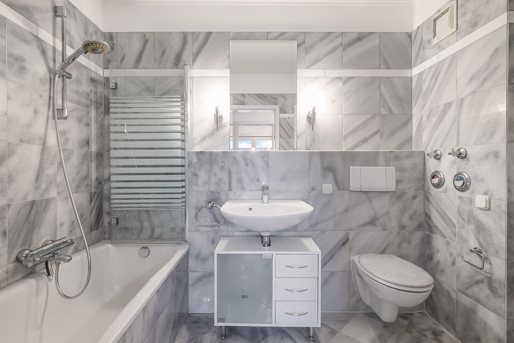 Modernes Badezimmer mit Badewanne, Dusche, Waschbecken, Spiegel und Wand-WC, alles in grauen Fliesen.