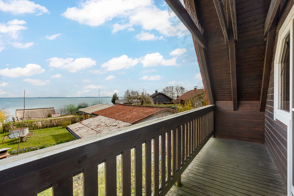 Holzbalkon mit Blick auf Gärten, Dächer und einen See im Hintergrund.
