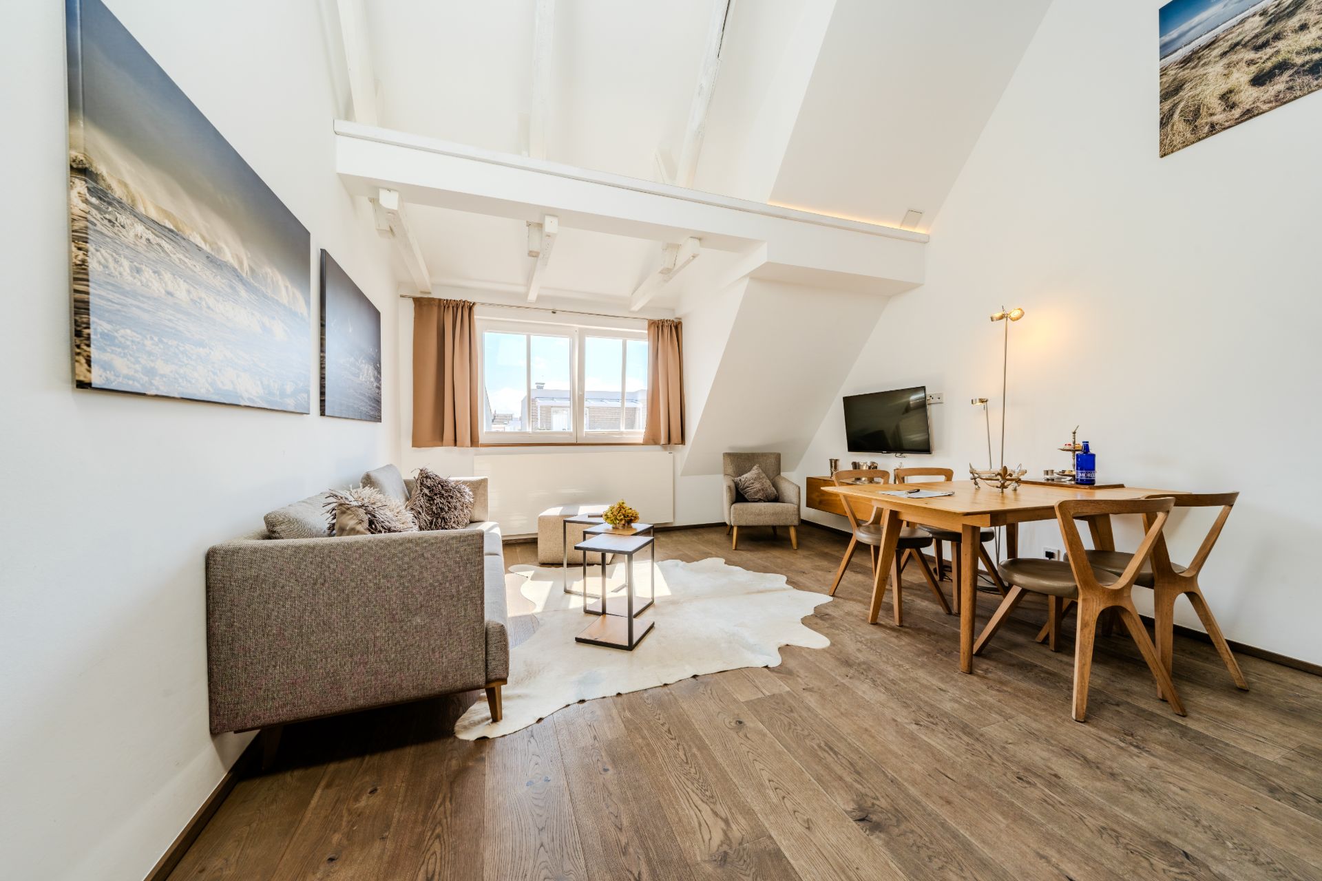 Helles Wohnzimmer mit Sofa, Esstisch, Fernseher und großen Fenstern, Holzboden und Dekoration an den Wänden.