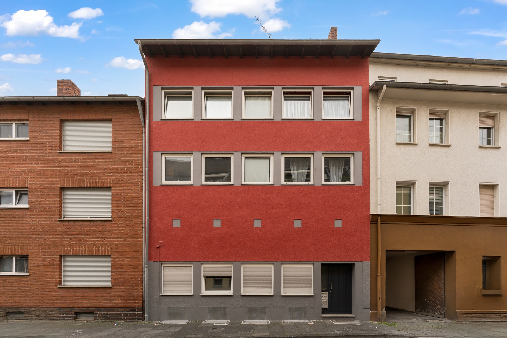 Dreistöckiges rotes Wohnhaus mit mehreren Fenstern und Tür, zwischen zwei anderen Gebäuden.