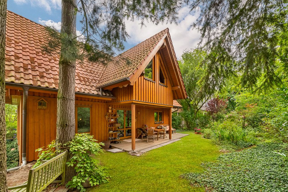 Holzhaus mit überdachter Terrasse und Garten, umgeben von Bäumen und viel Grün.