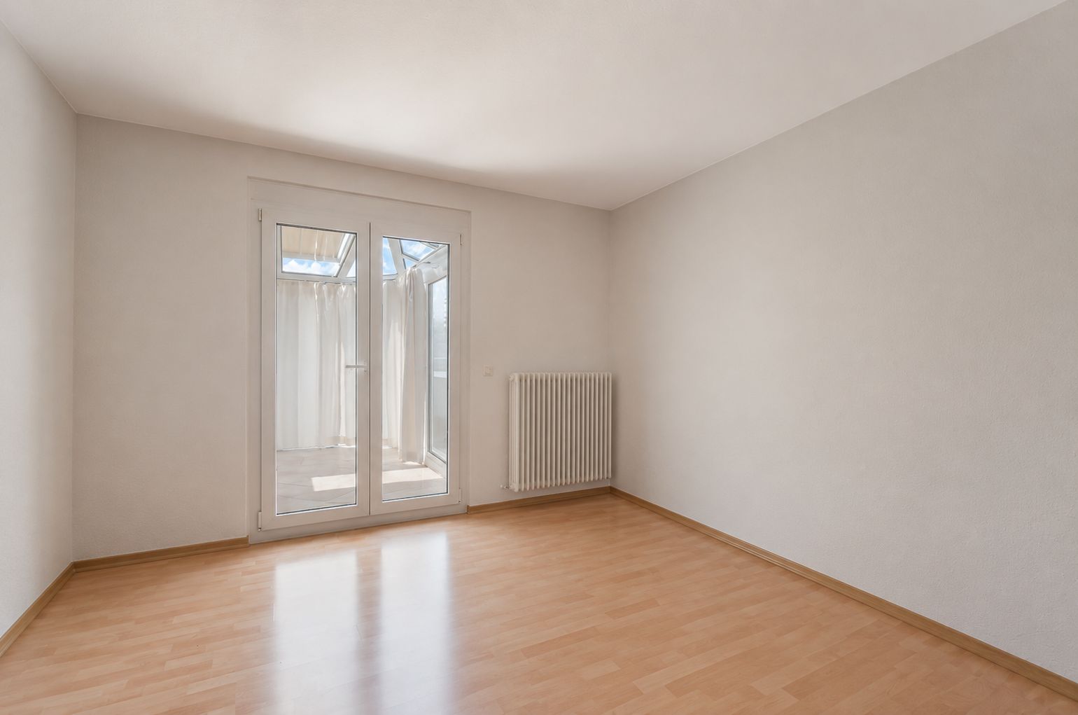 Leerer Raum mit hellem Holzboden, weißen Wänden und Glastür zu einem Balkon.