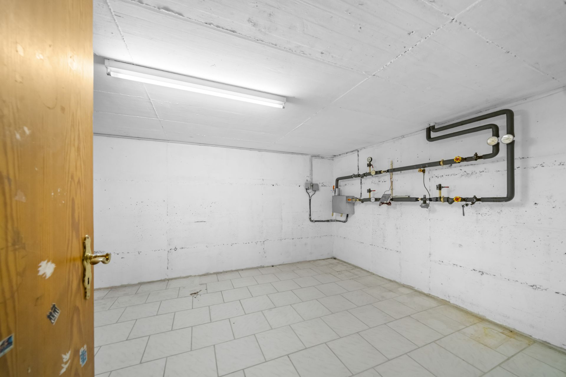 Leerer Kellerraum mit weißen Wänden, Fliesenboden und sichtbaren Rohren an der Wand.