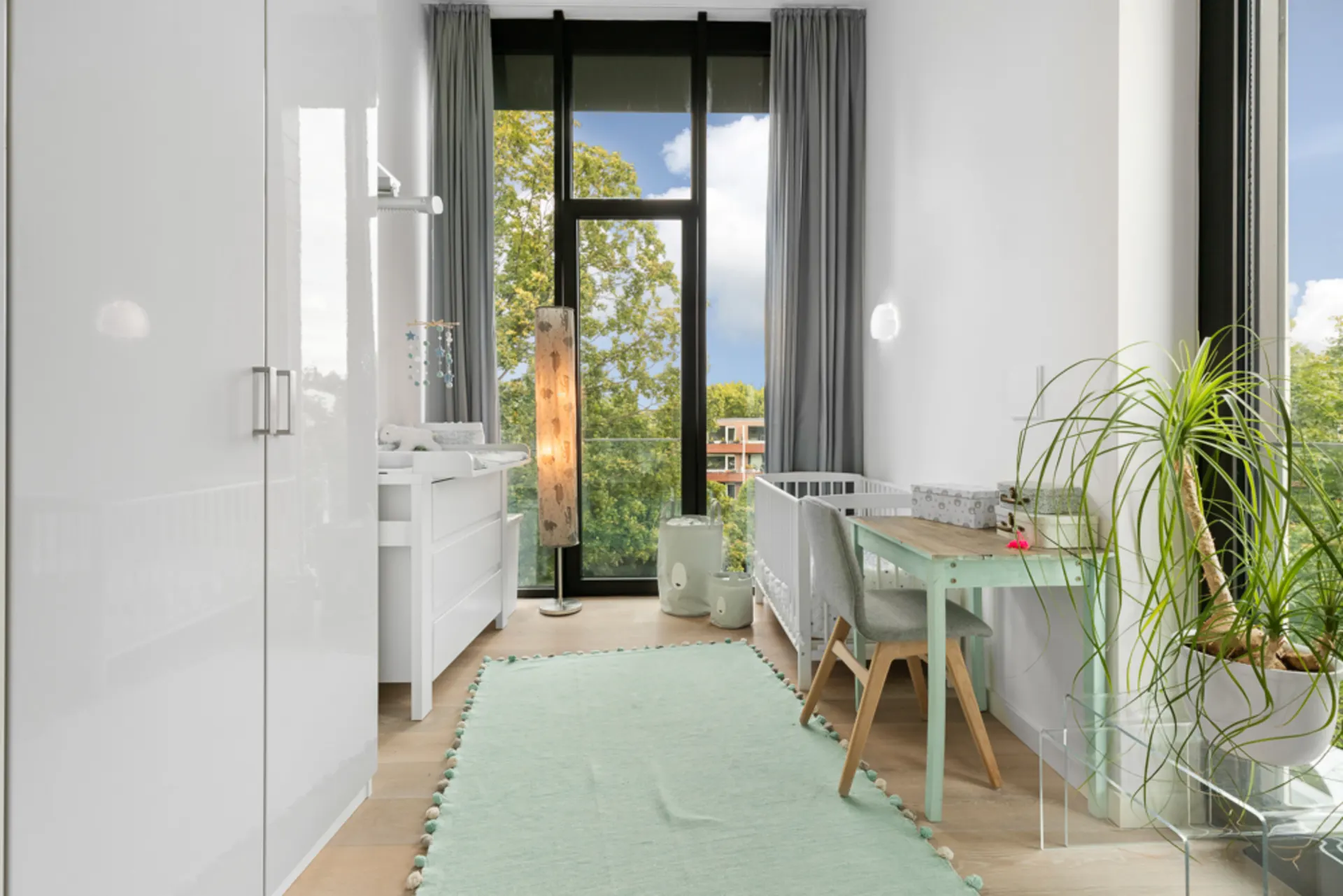 Helles Kinderzimmer mit großem Fenster, Wickeltisch, Schreibtisch, Kinderbett und Pflanze.