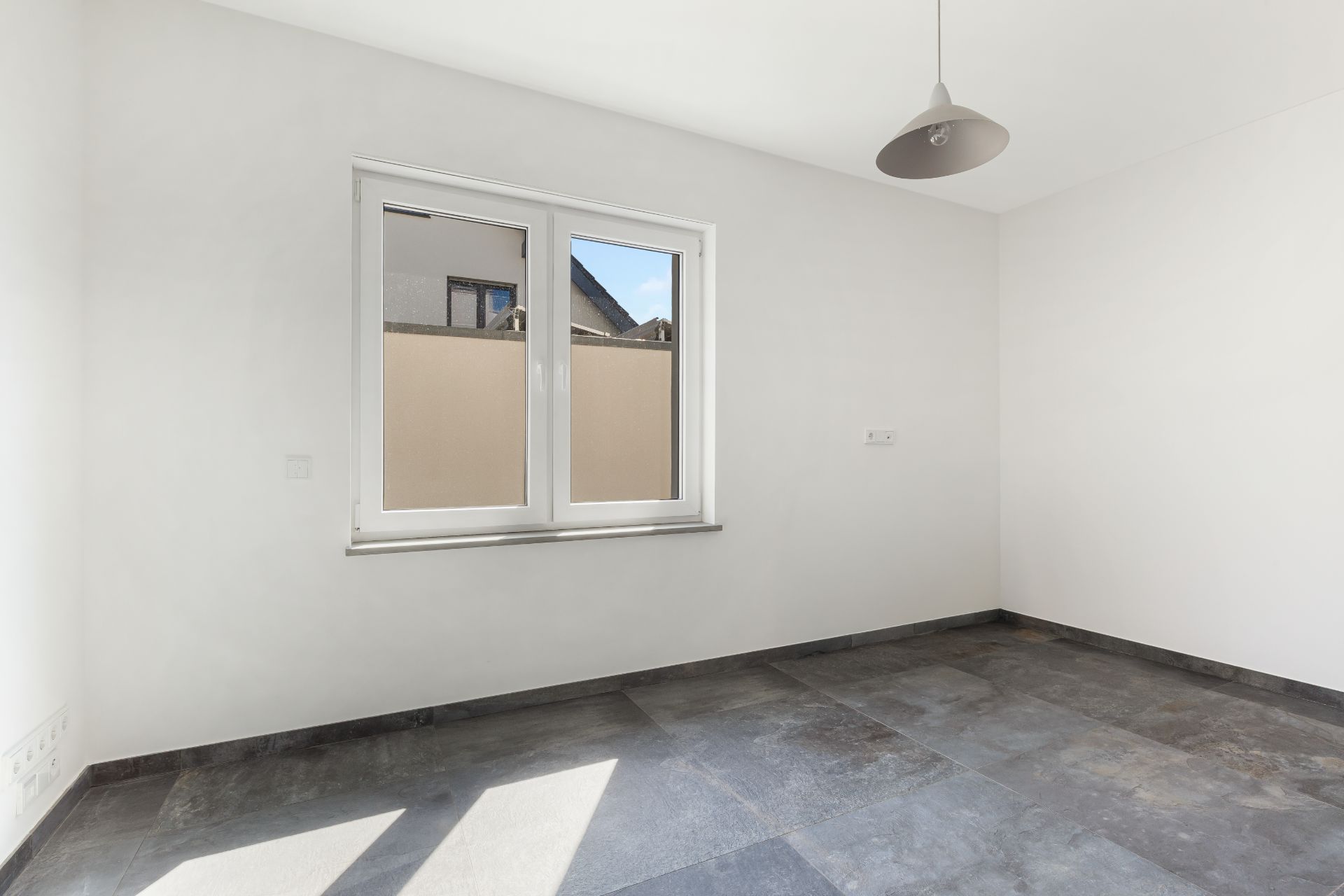 Leerer Raum mit grauem Fliesenboden, weißer Wand, Fenster und Deckenlampe.