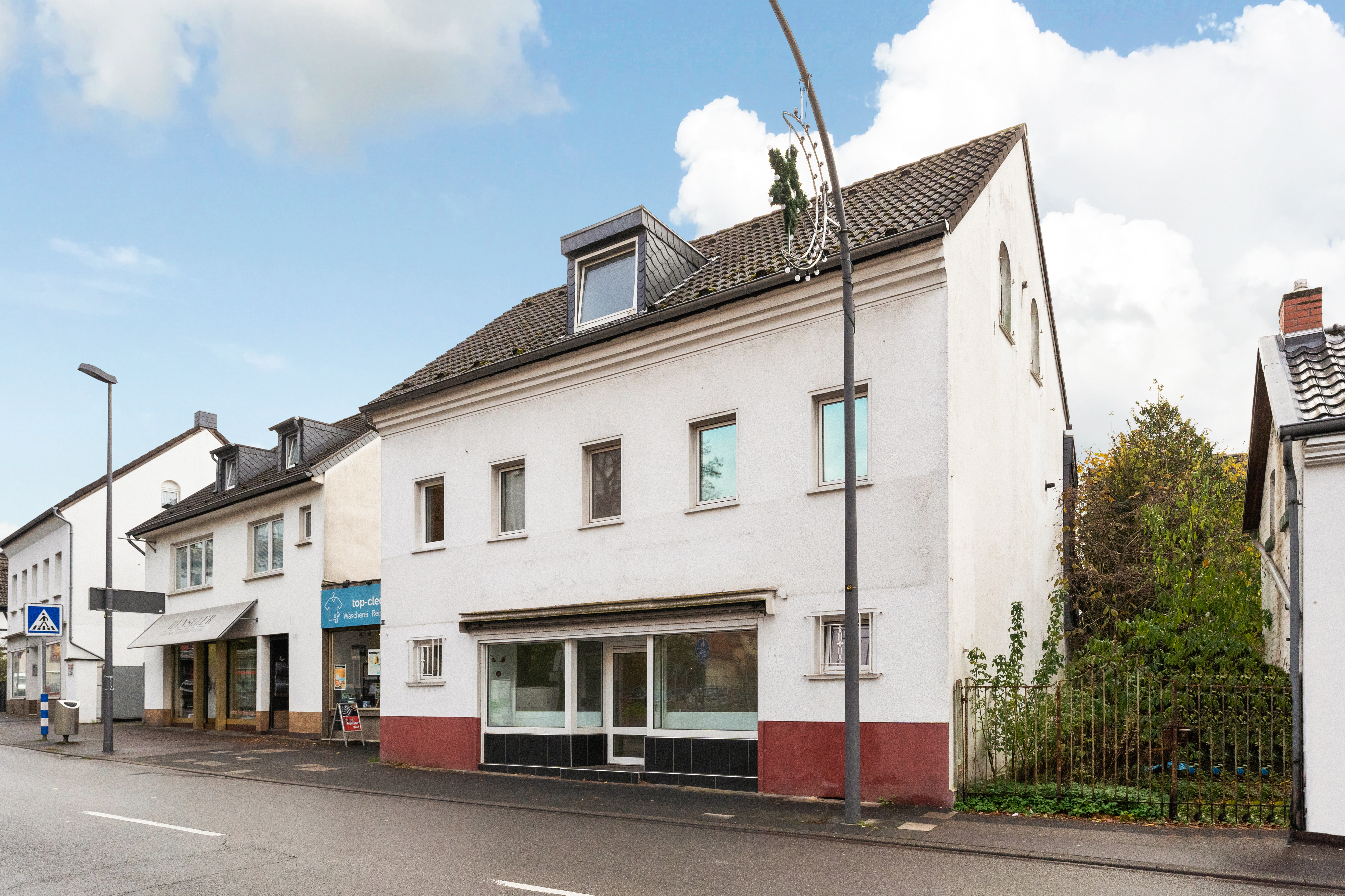 Weißes zweistöckiges Wohnhaus mit Schaufenster im Erdgeschoss an einer Straße.
