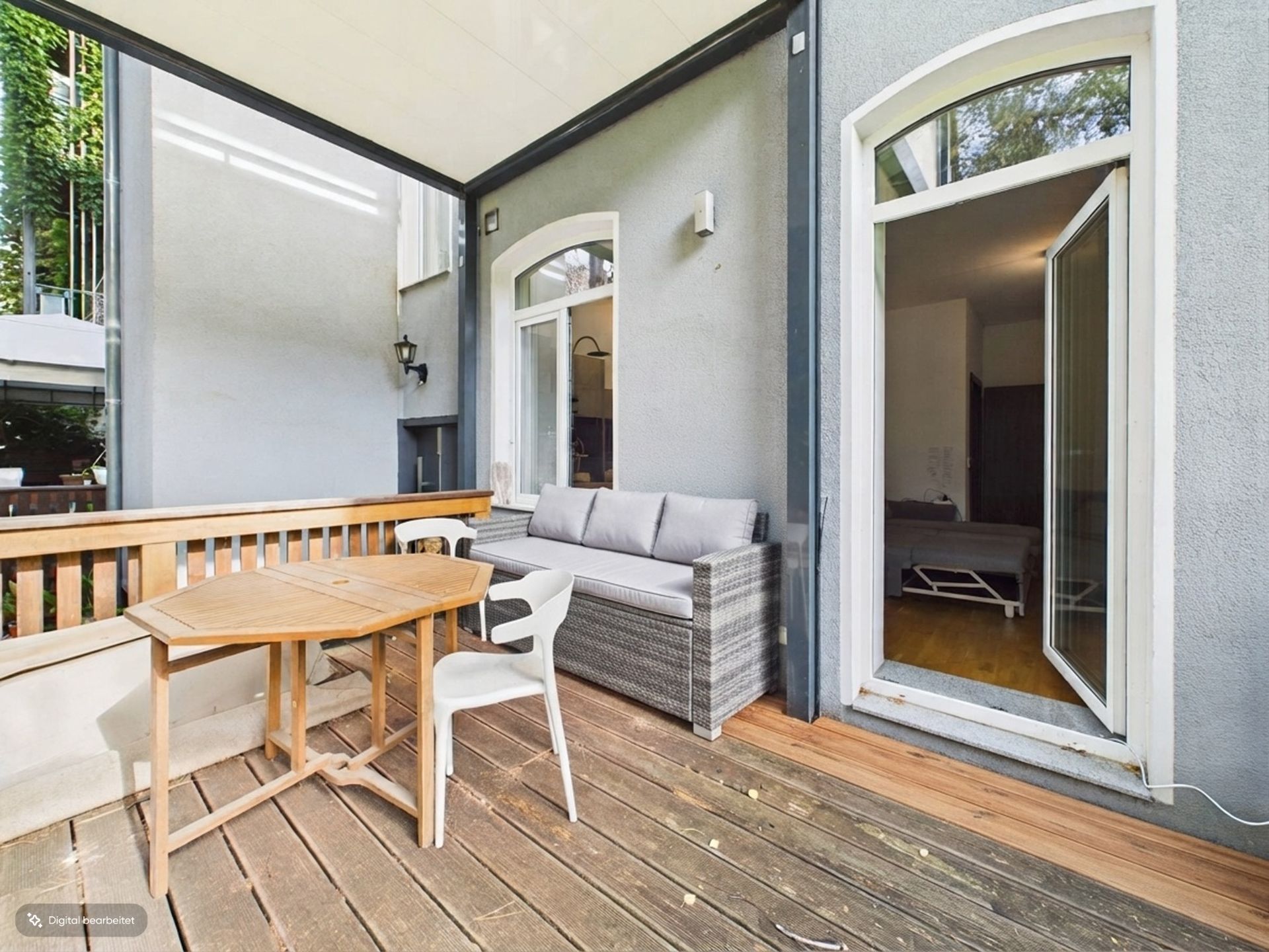 Überdachte Terrasse mit Holztisch, zwei Stühlen und grauem Sofa, Tür führt ins Schlafzimmer.