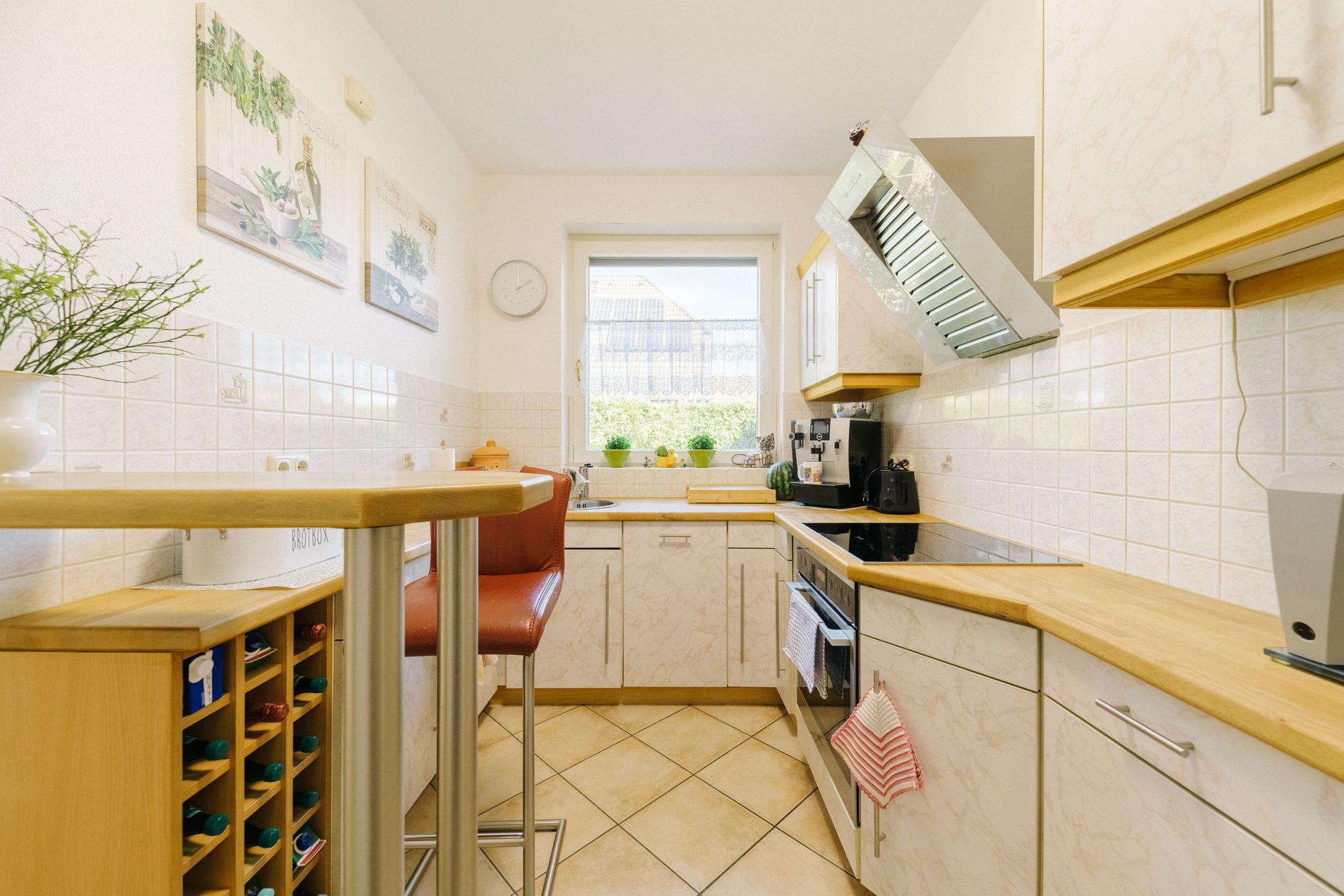 Helle Küche mit weißen Schränken, Holzakzenten, Esstheke, zwei Hockern und einem Fenster.