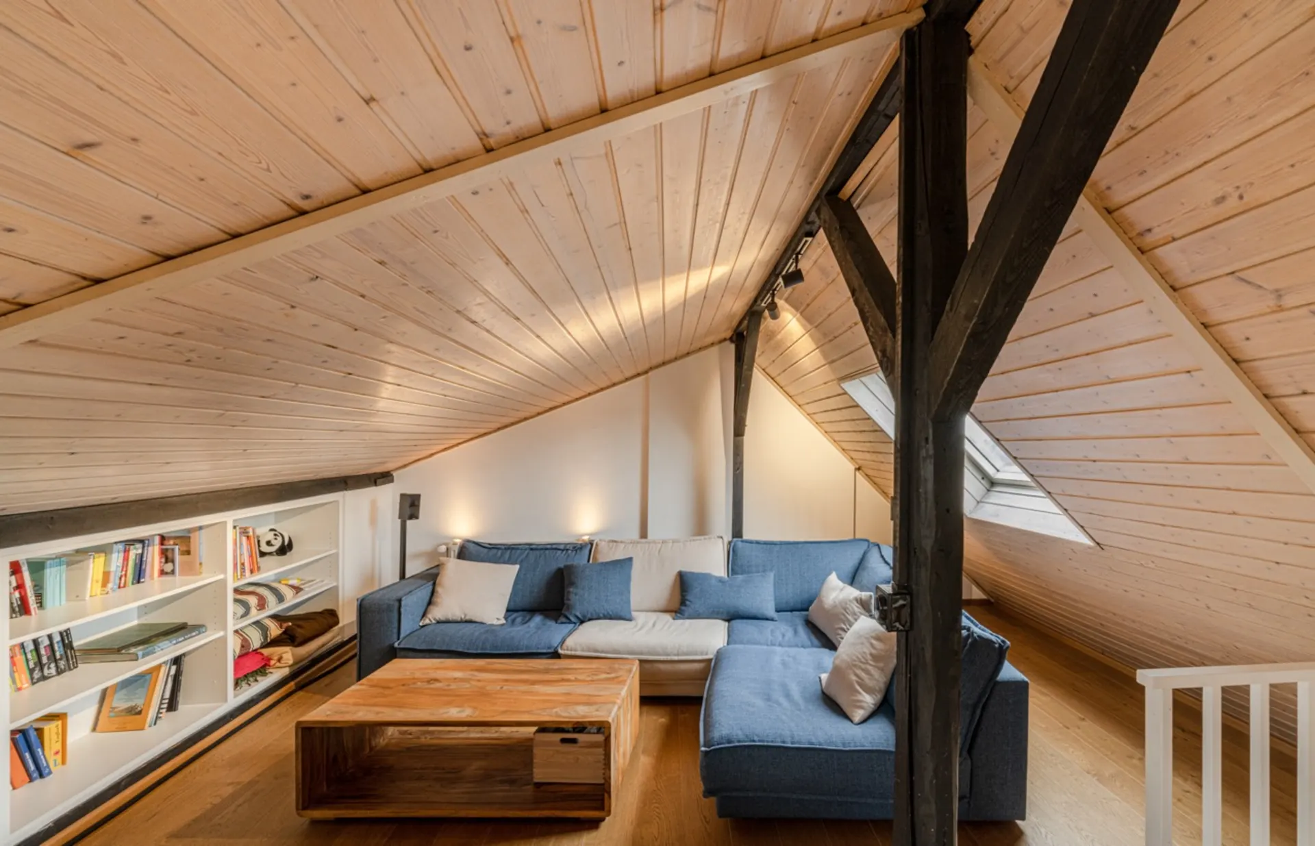 Gemütliches Dachgeschosszimmer mit blauer Eckcouch, Holzdecke und Bücherregal.