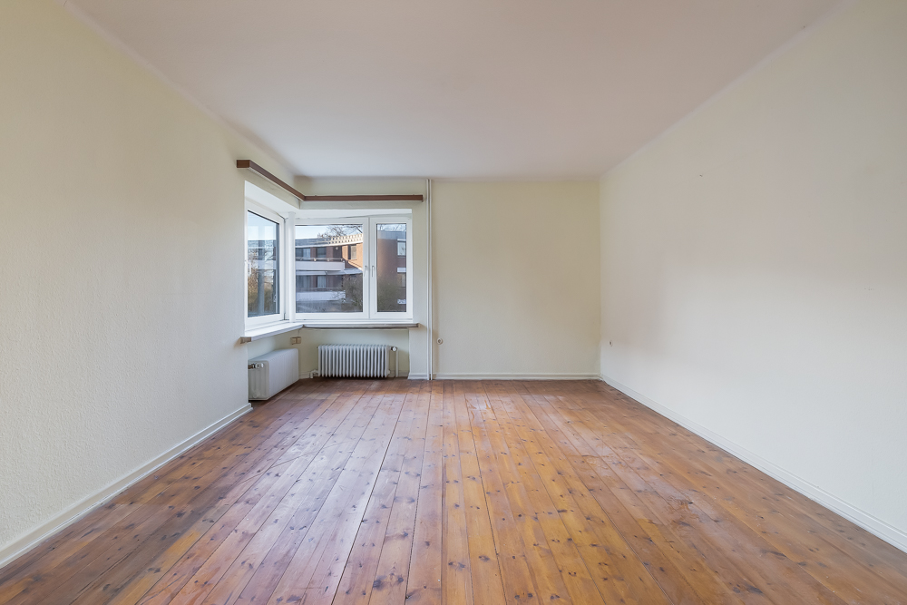 Leeres Zimmer mit Holzboden, großen Fenstern und Heizkörper unter dem Fenster.