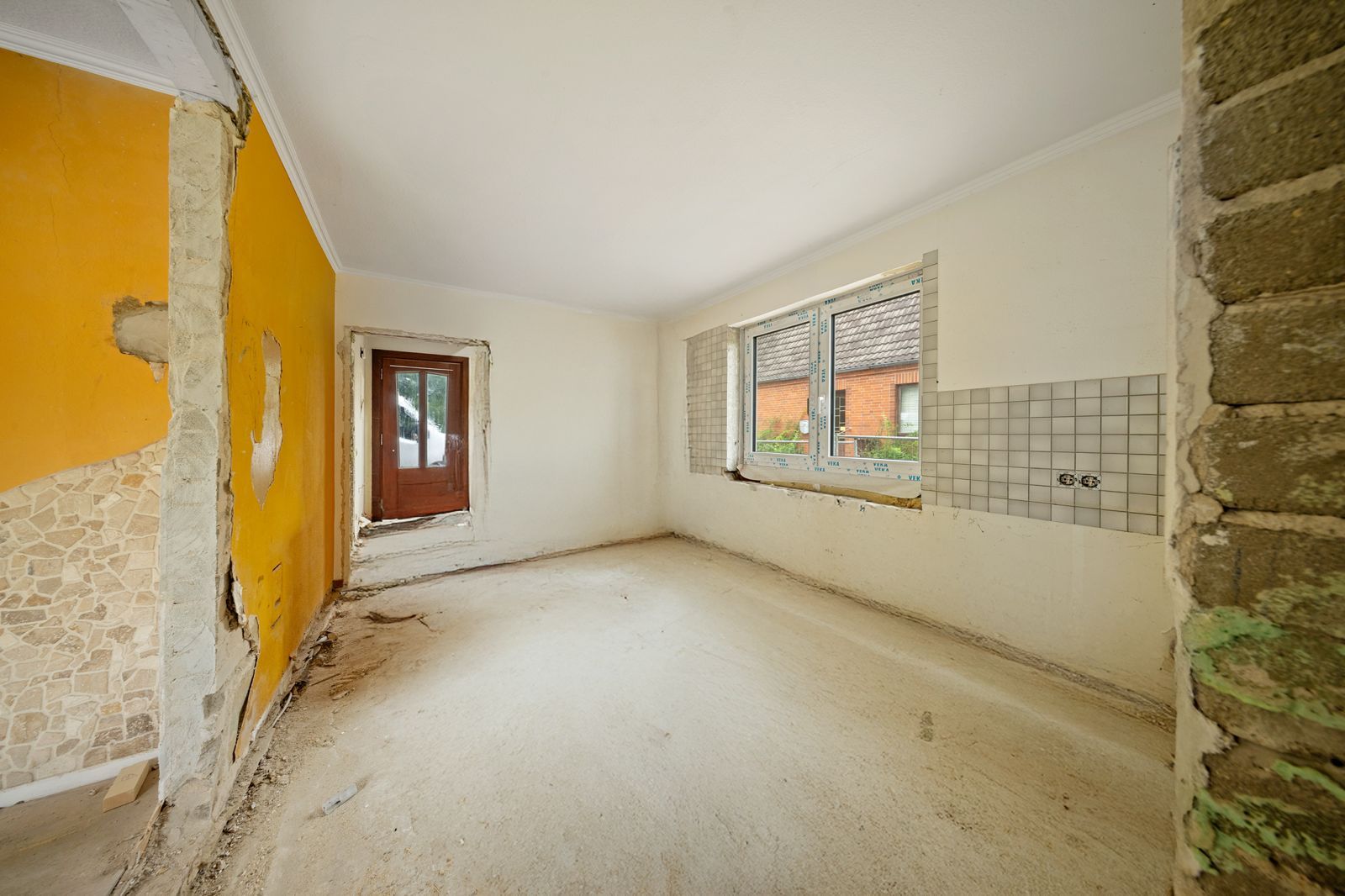 Leerer, renovierungsbedürftiger Raum mit gelber Wand, Fenster und Tür, teilweise freigelegte Wände.