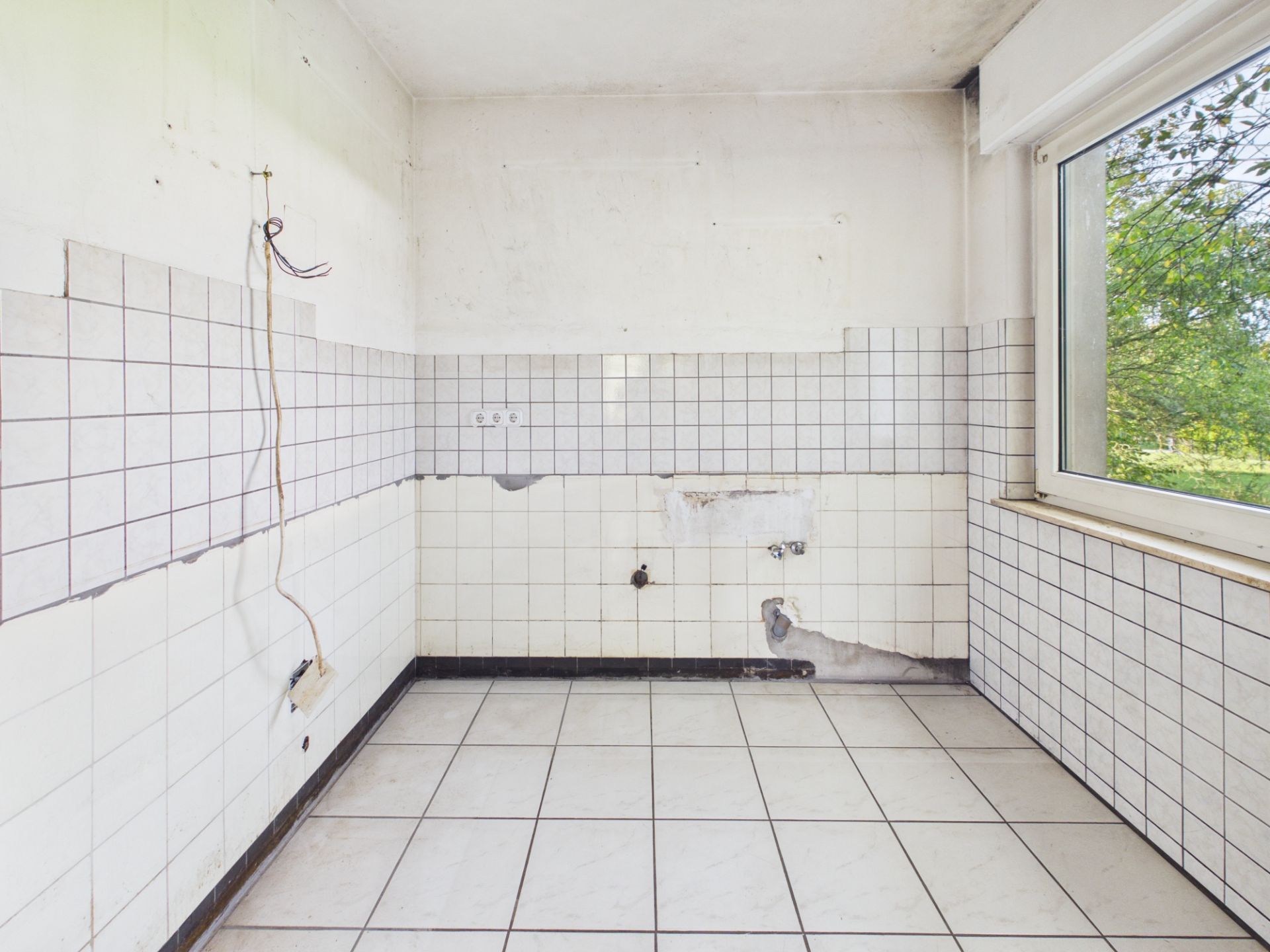 Leerer, gefliester Raum mit großem Fenster und sichtbaren Anschlüssen an den Wänden.