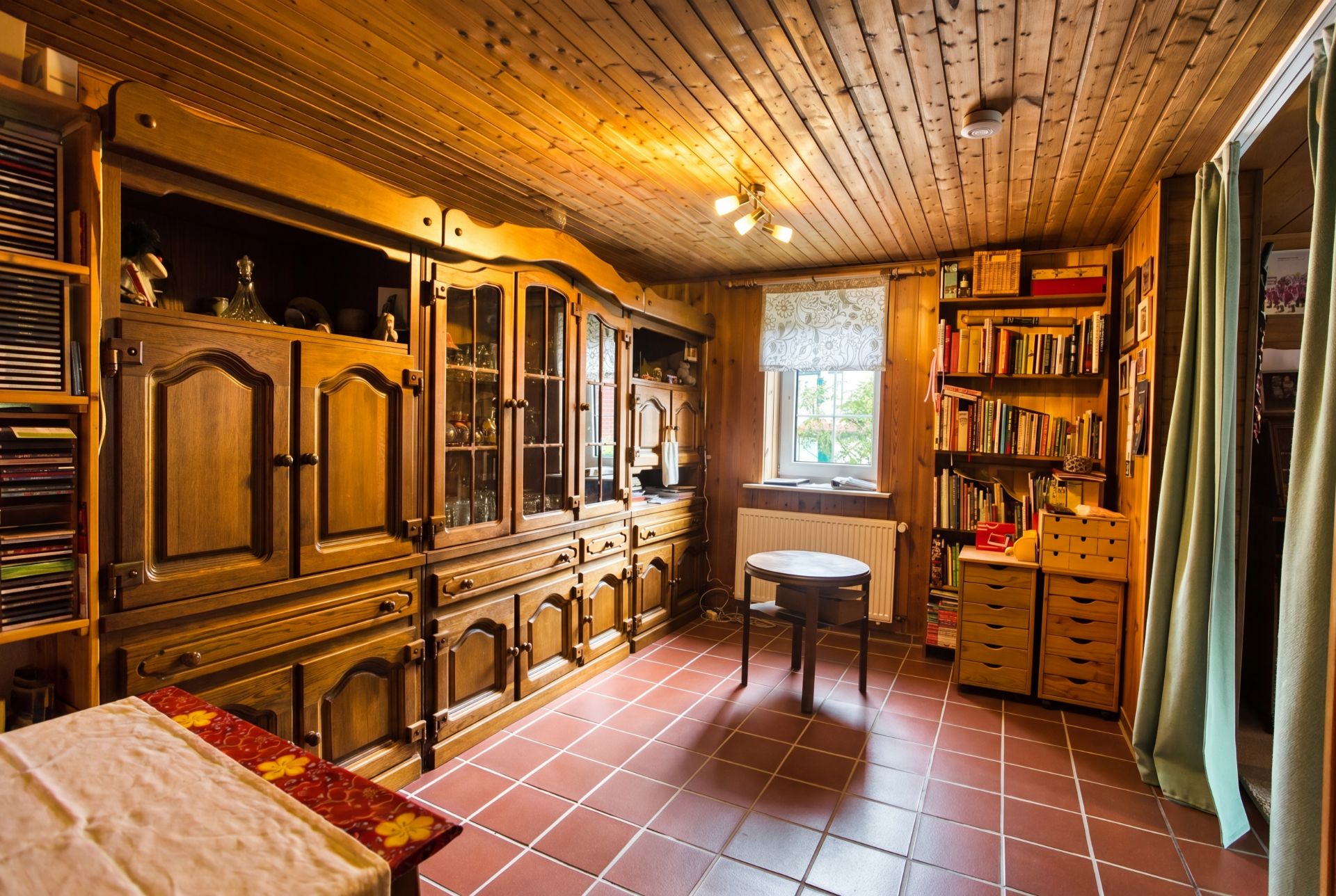 Wohnzimmer mit Holzmöbeln, Bücherregal, kleinem Tisch und Fenster, Fliesenboden und Holzdecke.