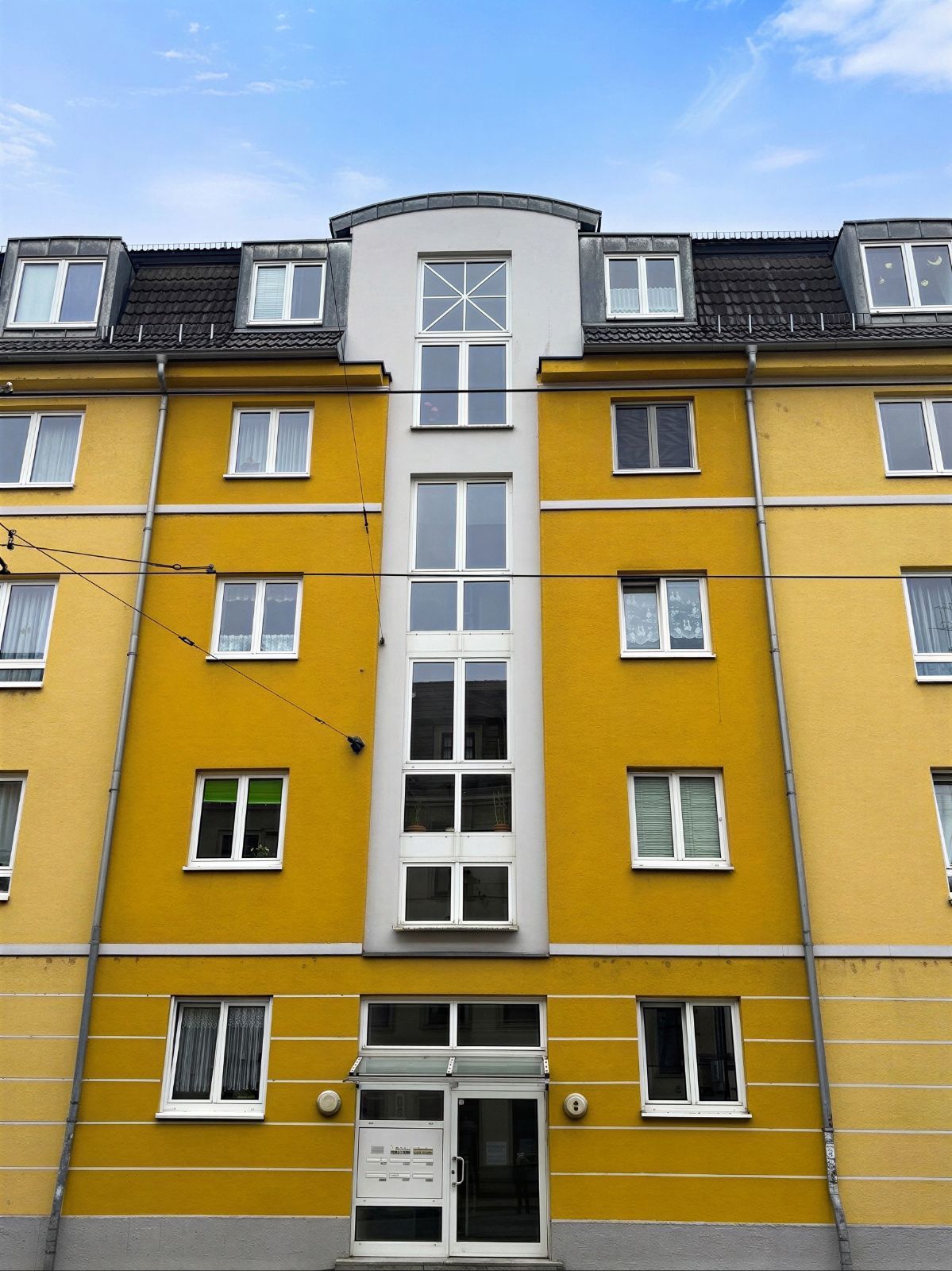 Gelbes Mehrfamilienhaus mit vielen Fenstern und Glaseingangstür, blauer Himmel darüber.