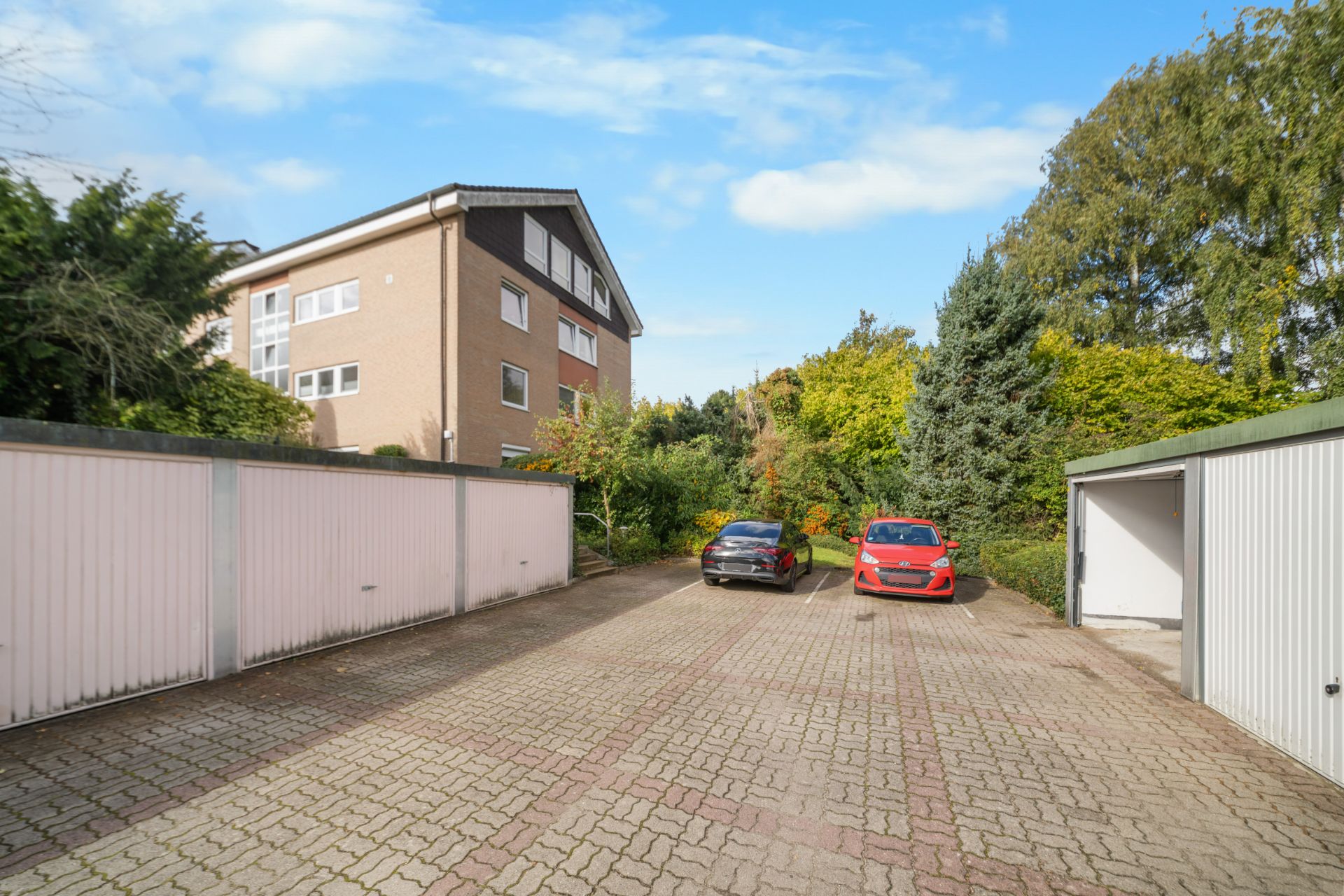Gepflasterter Parkplatz mit zwei Autos, Garagen und einem Wohngebäude im Hintergrund.
