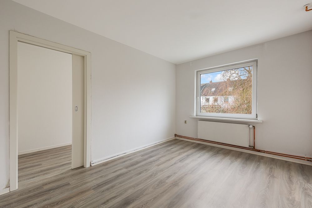 Leeres Zimmer mit großem Fenster, heller Wand und Laminatboden, verbindet mit weiterem Raum.