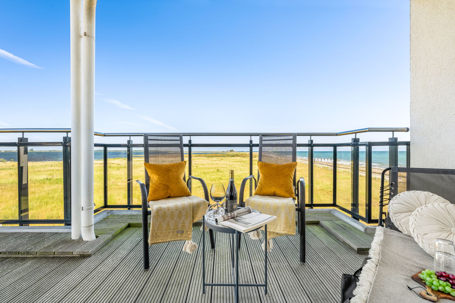 Balkon mit zwei Stühlen, Tisch, Kissen und Ausblick auf Wiese und Meer.
