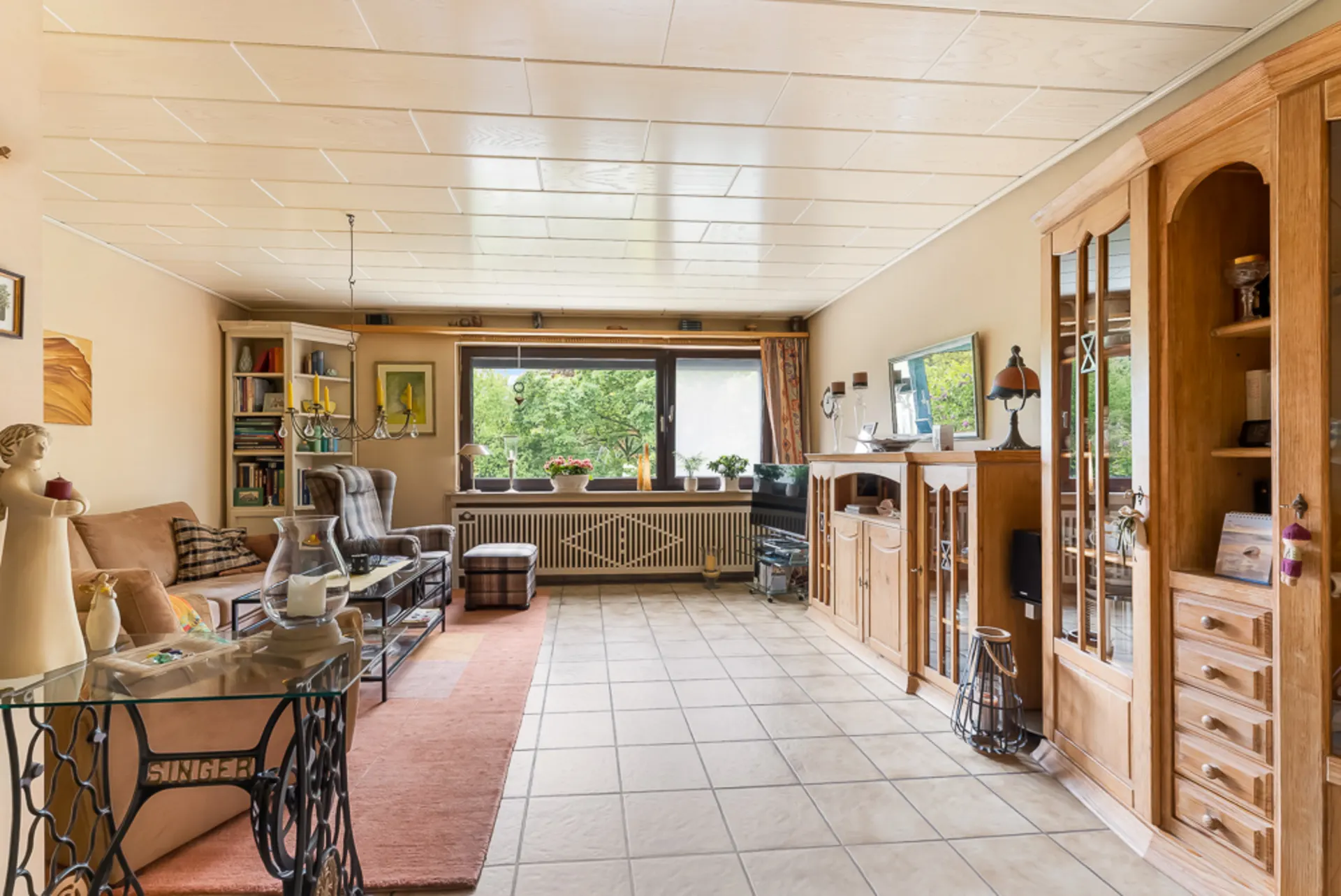 Helles Wohnzimmer mit Fliesenboden, Holzmöbeln, Sofa, Sessel und großem Fenster mit Blick ins Grüne.