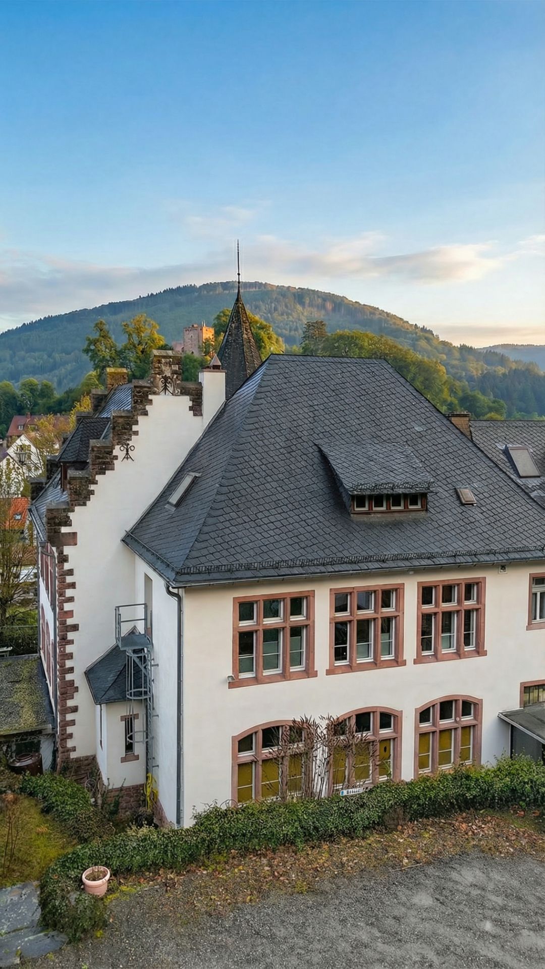 Großes, weißes Wohnhaus mit vielen Fenstern und dunklem Dach, im Hintergrund bewaldete Hügel.