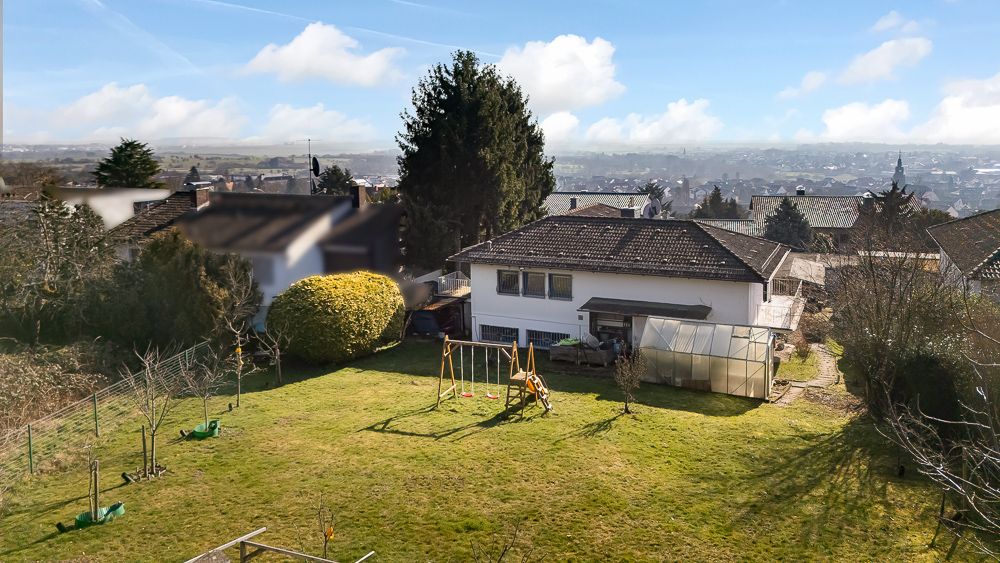 Haus mit großem Garten, Spielplatz und Gewächshaus an einem sonnigen Tag.