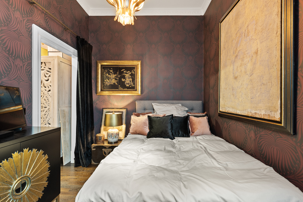 Kleines Schlafzimmer mit Doppelbett, dekorativen Kissen, Wandkunst und dunkler Tapete.