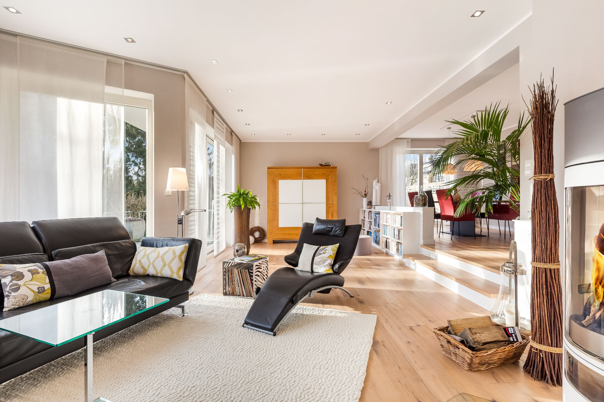 Helles Wohnzimmer mit schwarzen Sofas, Kamin, Pflanzen und großen Fenstern.