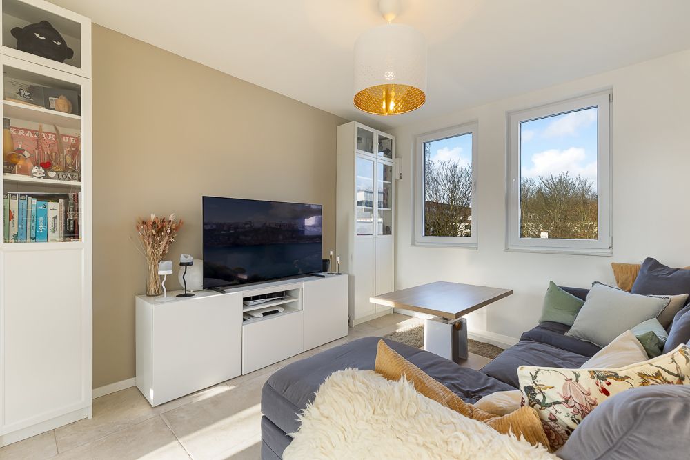 Helles Wohnzimmer mit Sofa, Couchtisch, Fernseher und Regal vor zwei Fenstern.