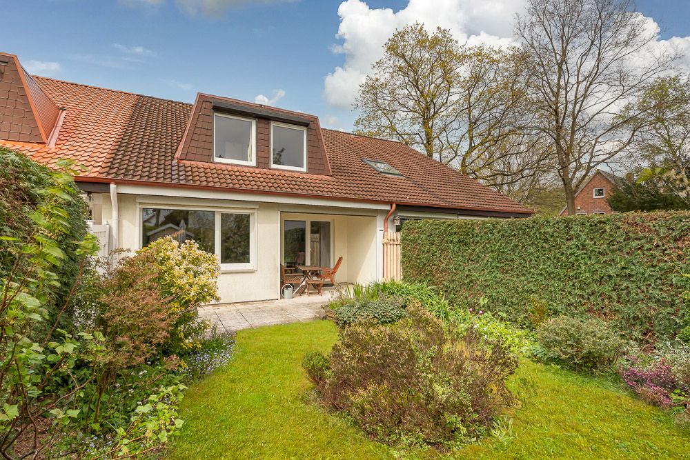 Haus mit Gartenterrasse, Rasenfläche und Hecke, umgeben von Büschen und Bäumen.
