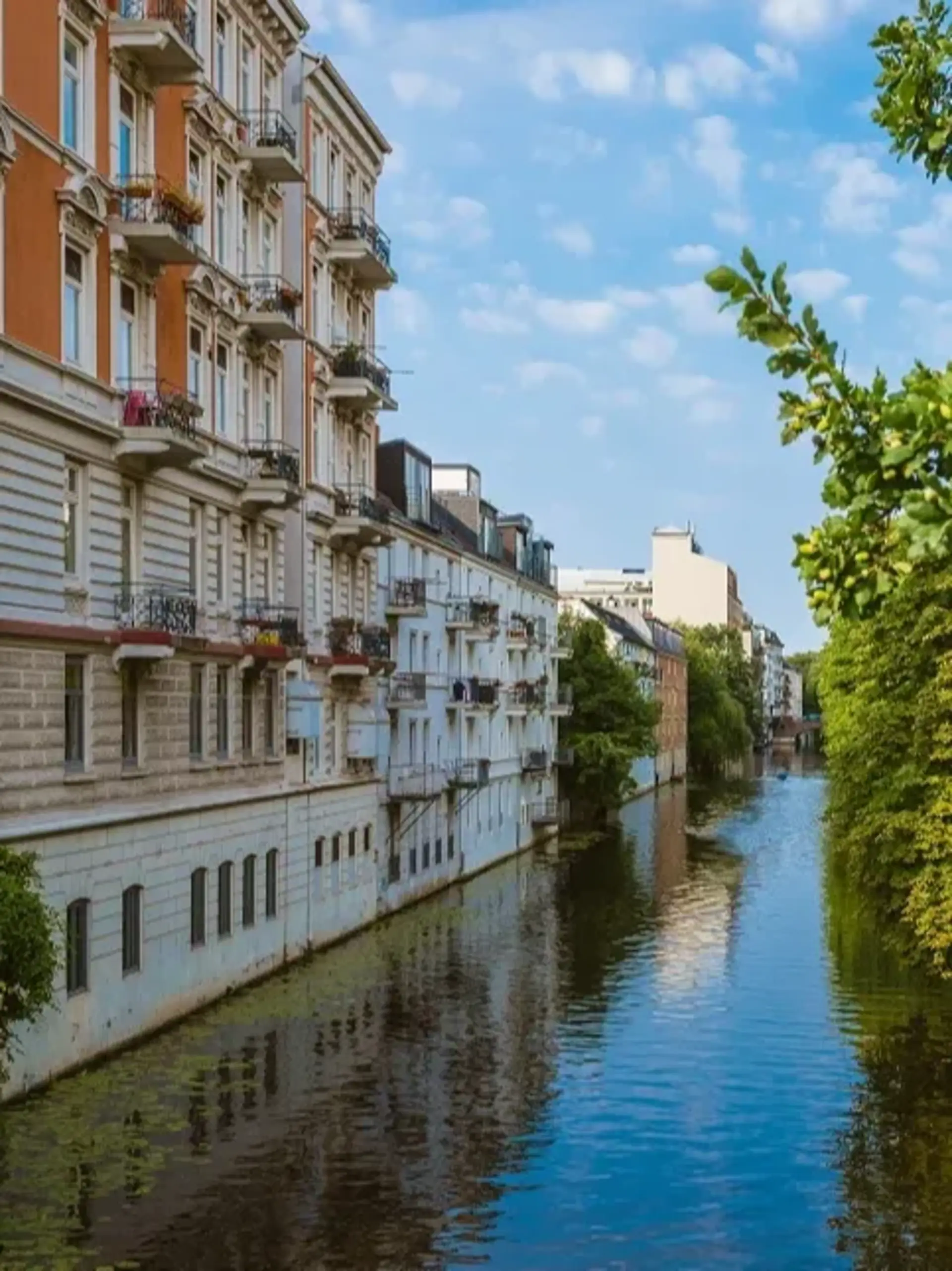 Wohngebäude mit Balkonen an einem Kanal, blauer Himmel und Bäume am Wasser.