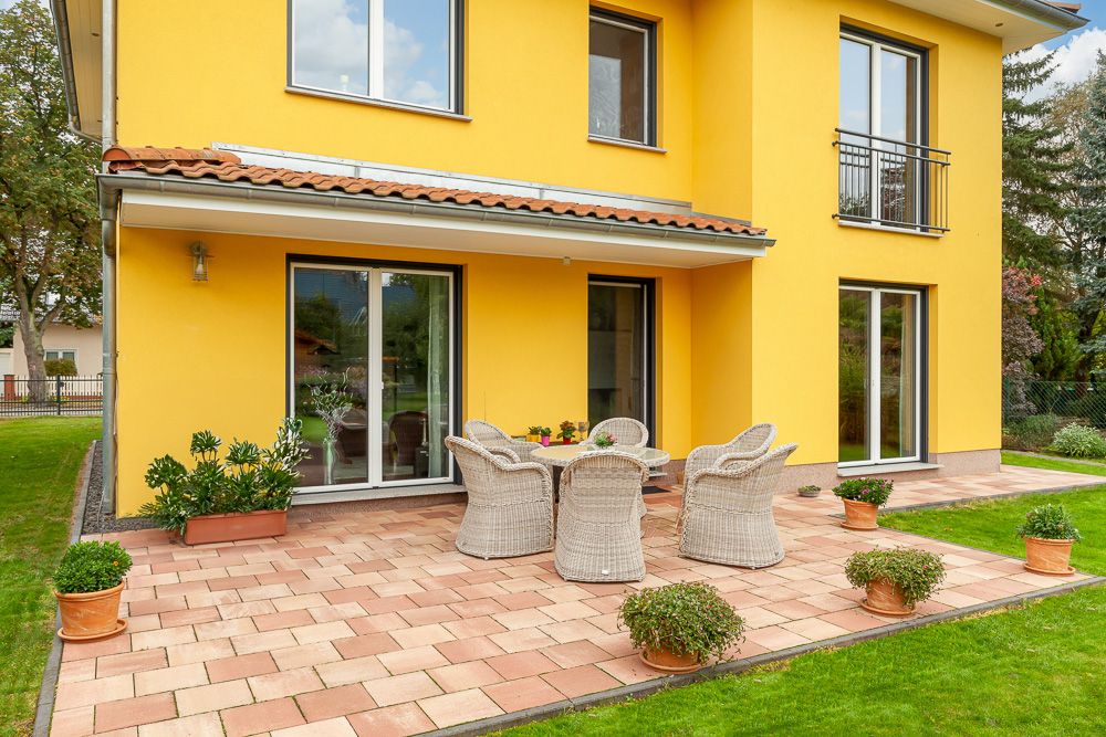 Gelbes Wohnhaus mit Terrasse, Rattanmöbeln und mehreren Pflanztöpfen auf den Fliesen.