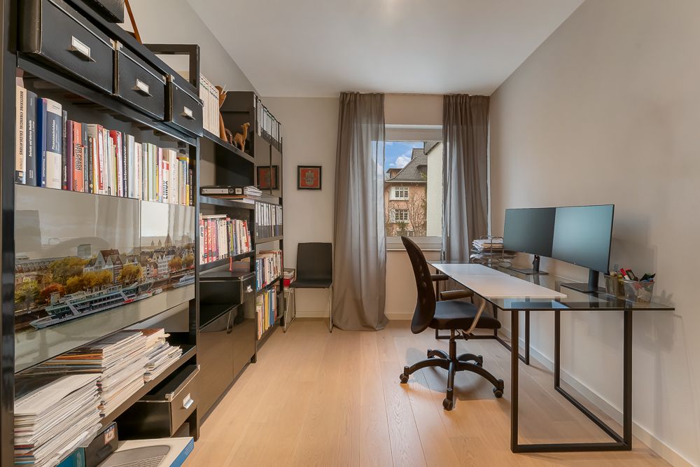 Helles Arbeitszimmer mit Schreibtisch, zwei Monitoren, Bücherregalen und Fenster mit Vorhängen.