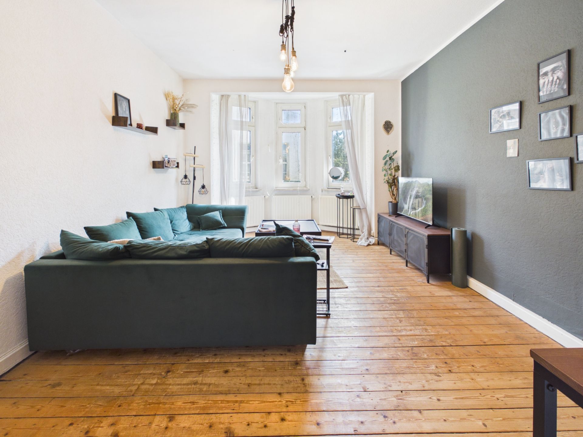 Wohnzimmer mit grünem Sofa, Holzboden, TV, Wandbildern und großen Fenstern mit weißen Vorhängen.