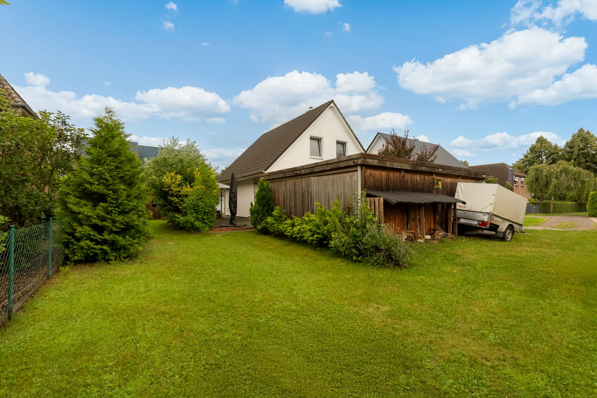 Weißes Einfamilienhaus mit Garten, umgeben von Bäumen und Rasen, Holzschuppen und Anhänger rechts.