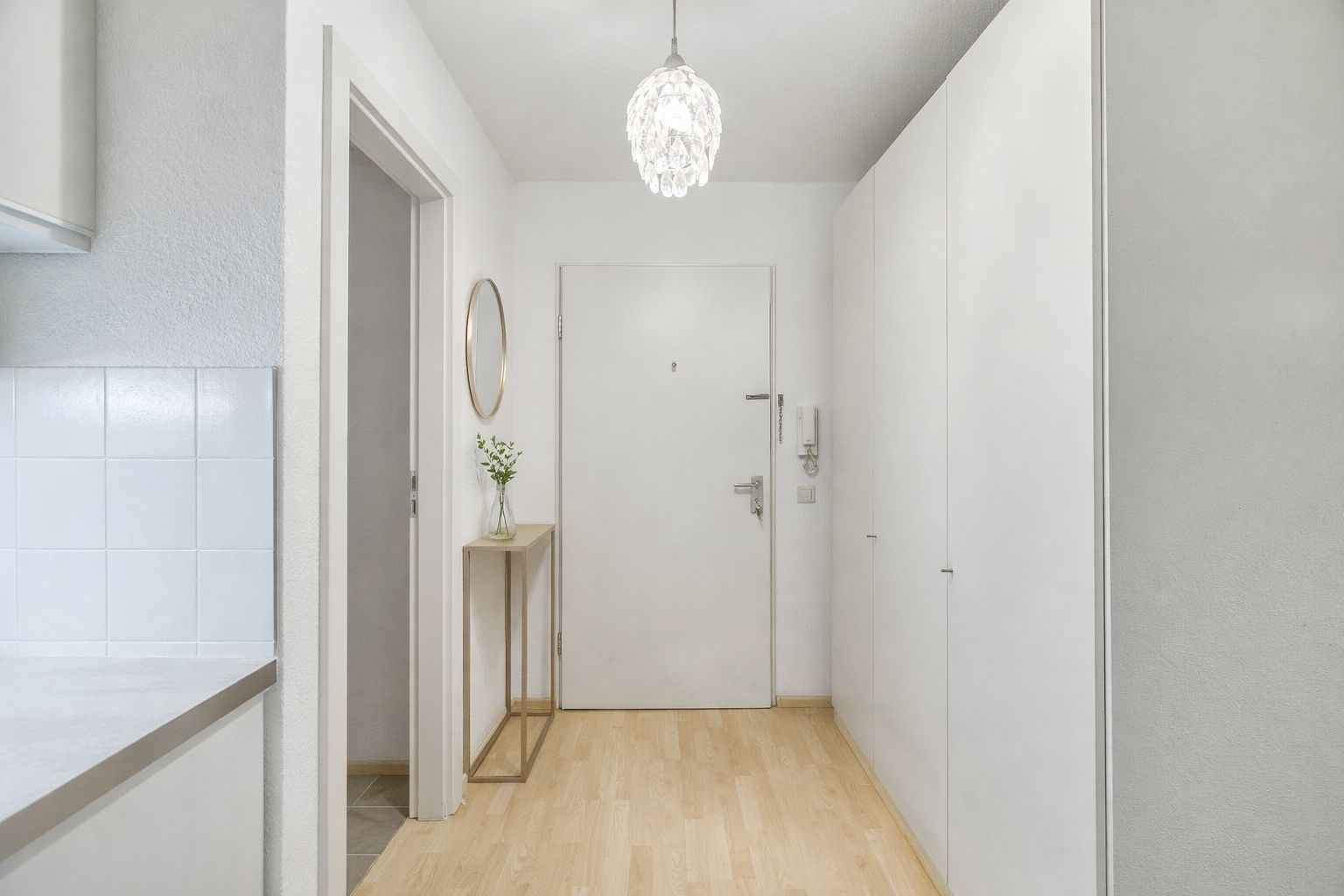 Heller Flur mit Holzboden, weißer Tür, Spiegel und kleinem Konsolentisch mit Vase.