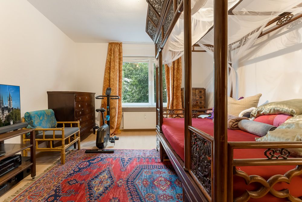 Schlafzimmer mit Himmelbett, Teppich, Heimtrainer, TV, Sessel und Kommoden vor Fenster.