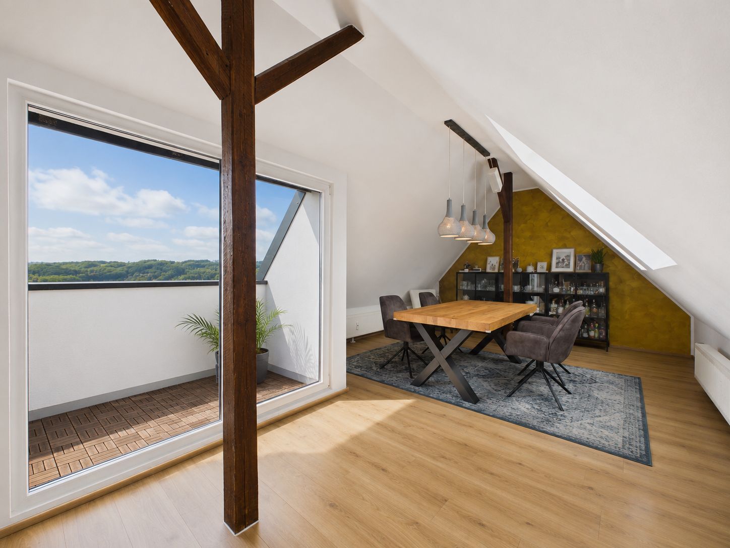Helles Dachgeschosszimmer mit Essbereich, Holztisch, Teppich und Zugang zu einem Balkon.