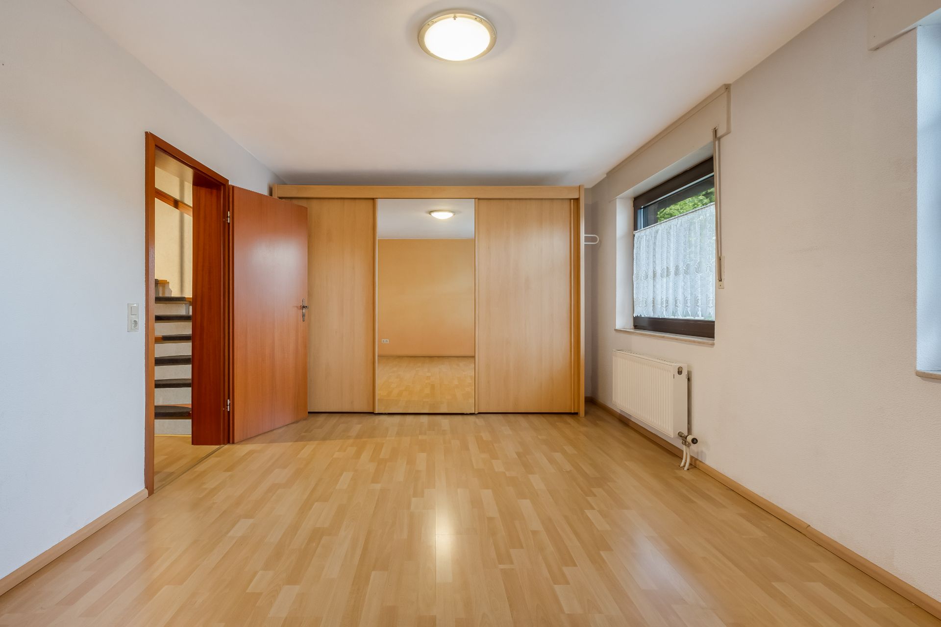 Leerer Raum mit Holzfußboden, großem Schrank mit Spiegel und Fenster mit Heizkörper.