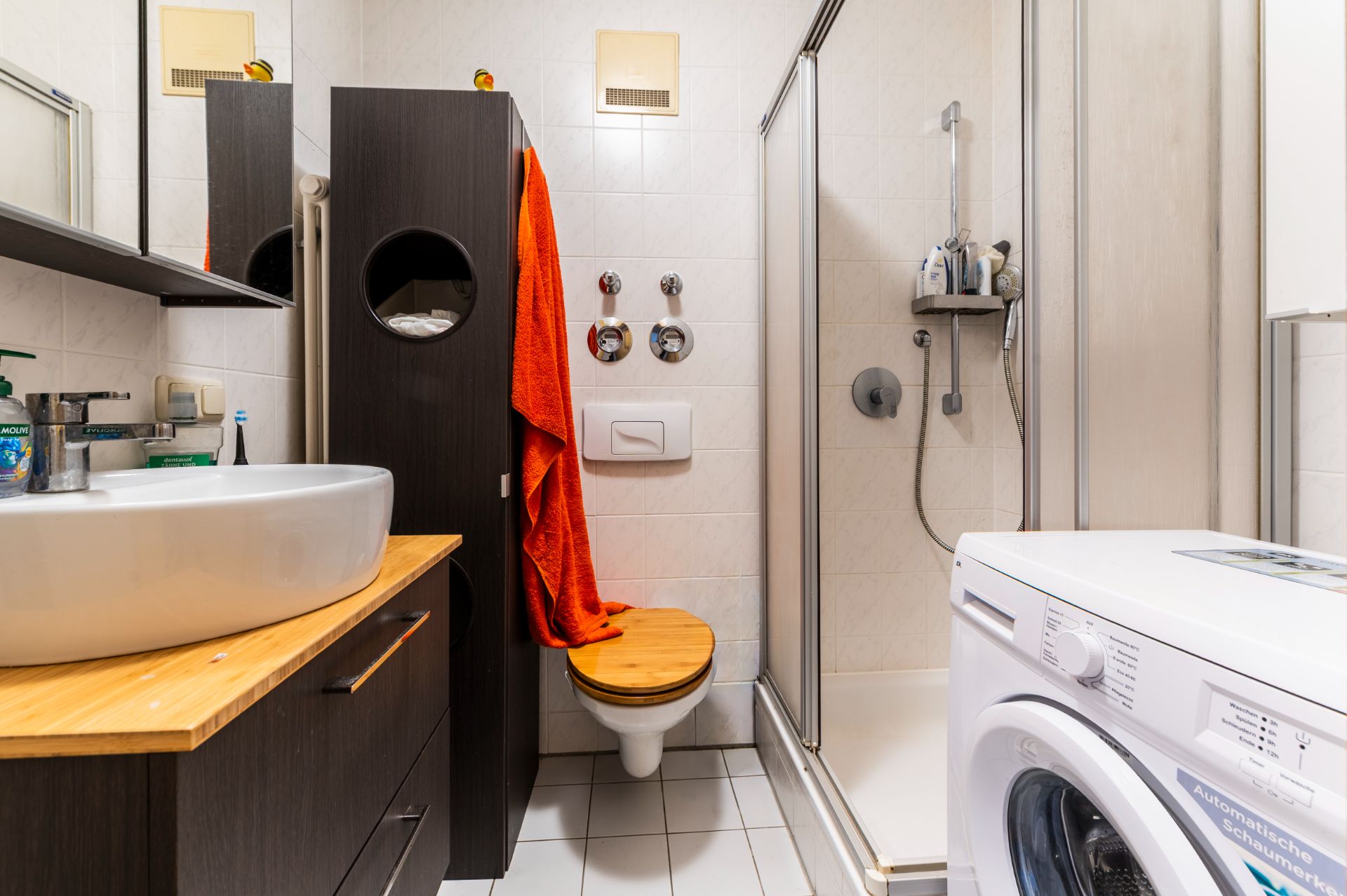 Kleines Badezimmer mit Dusche, Waschbecken, WC, Waschmaschine und dunklen Möbeln.