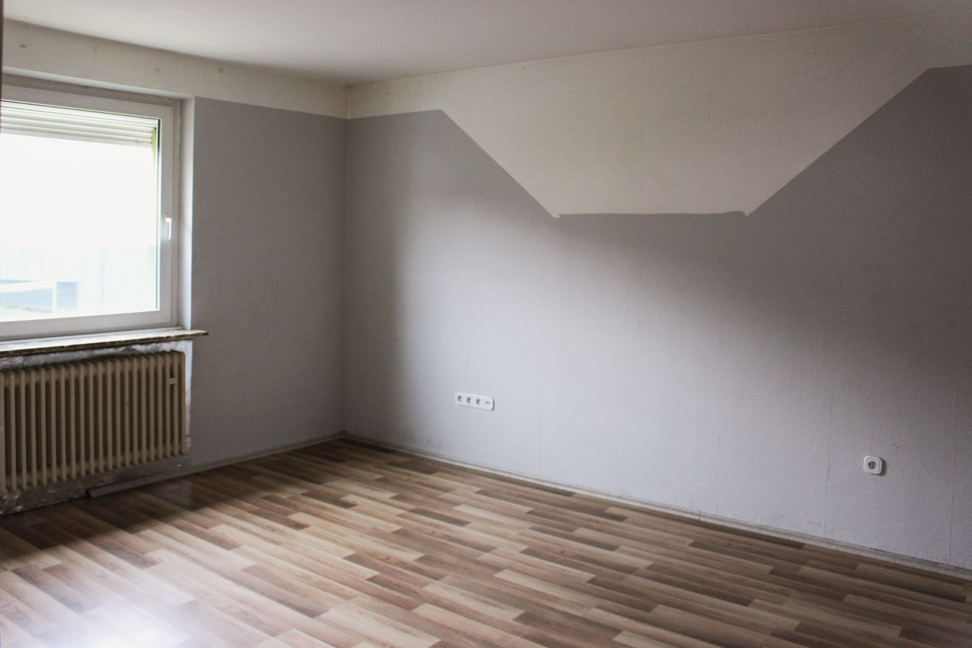 Leerer Raum mit hellem Laminatboden, grauen Wänden und einem Fenster mit Heizkörper.