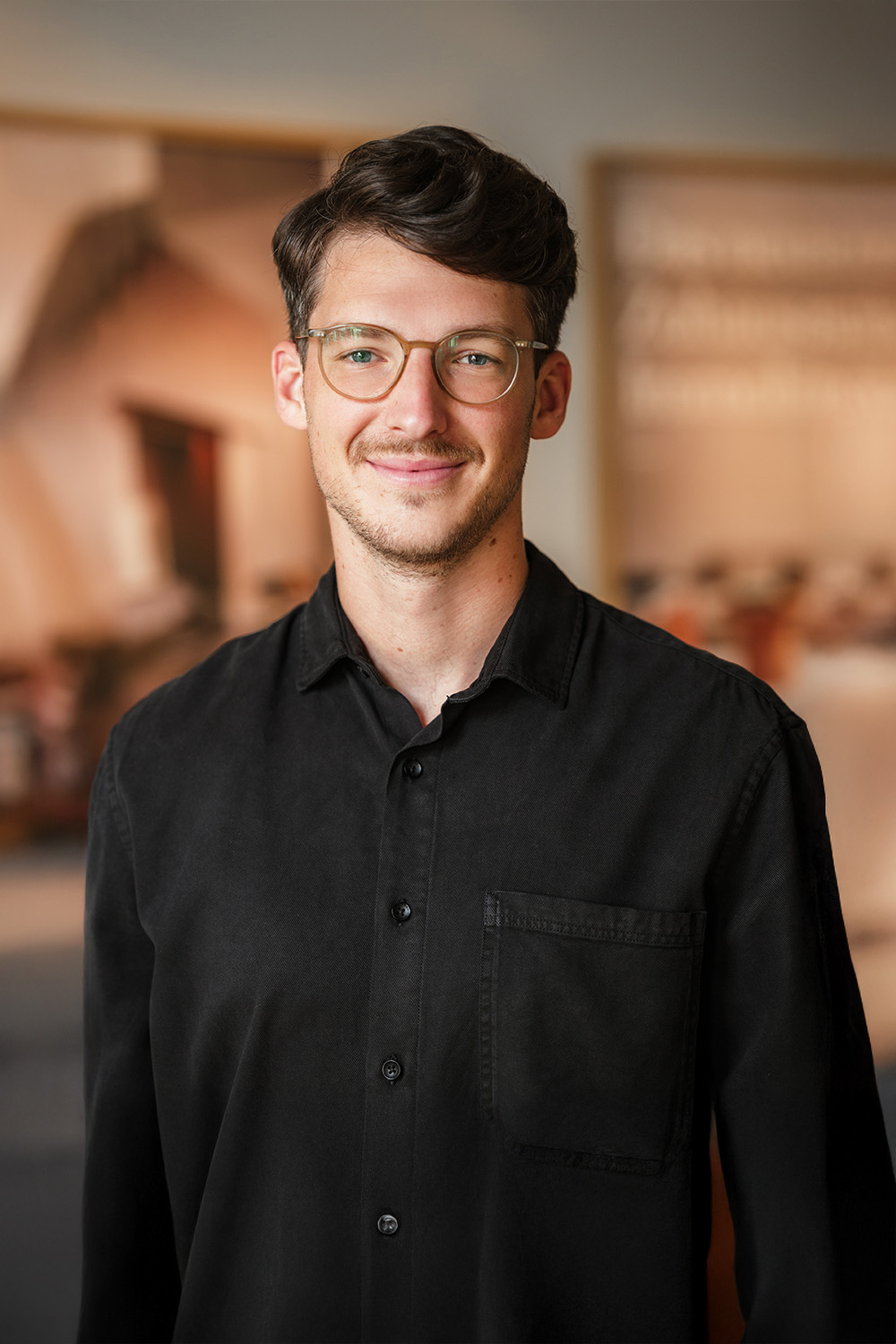 Ein junger Mann mit Brille und schwarzem Hemd steht in einem Innenraum und lächelt in die Kamera.