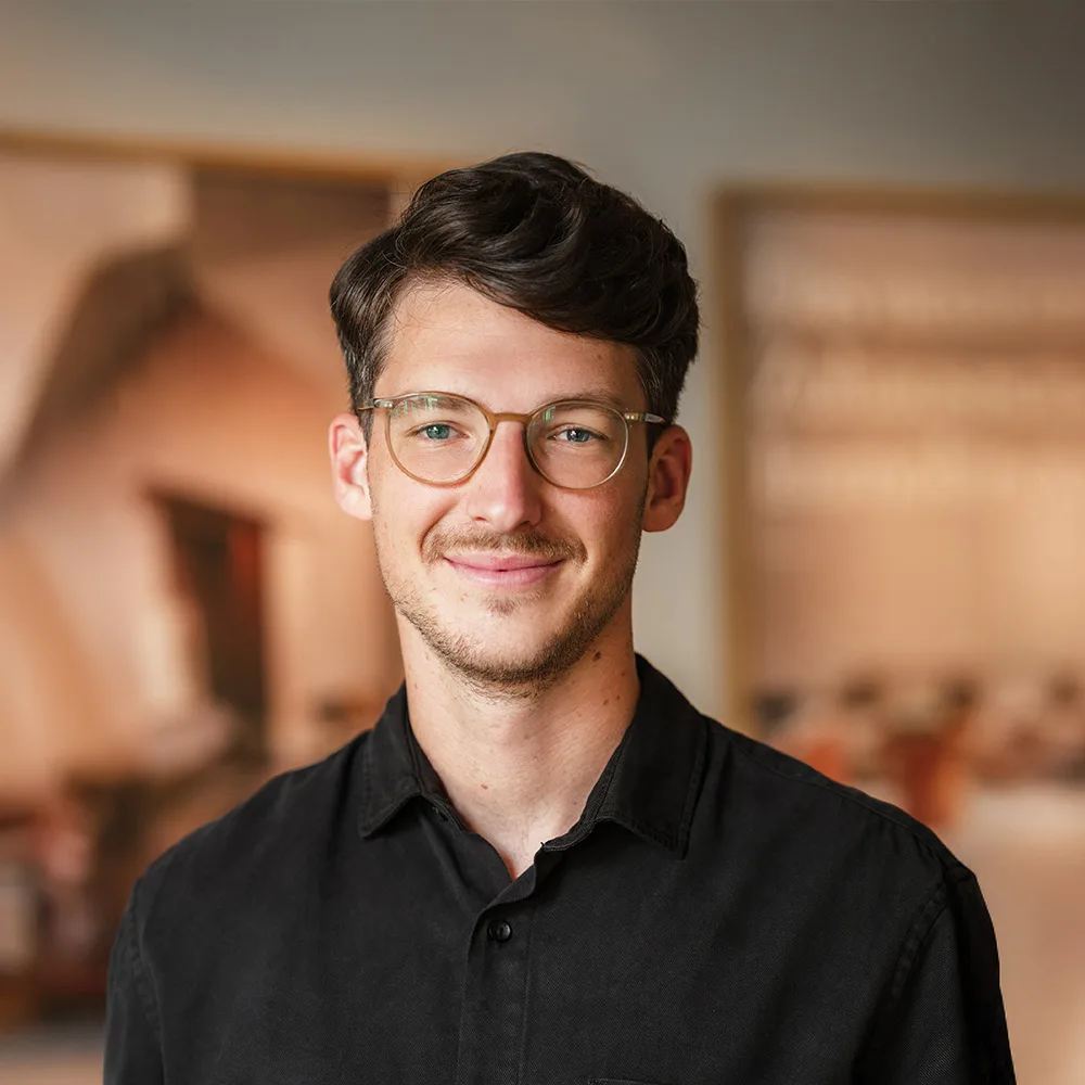 Ein junger Mann mit Brille und schwarzem Hemd steht in einem Innenraum und lächelt in die Kamera.