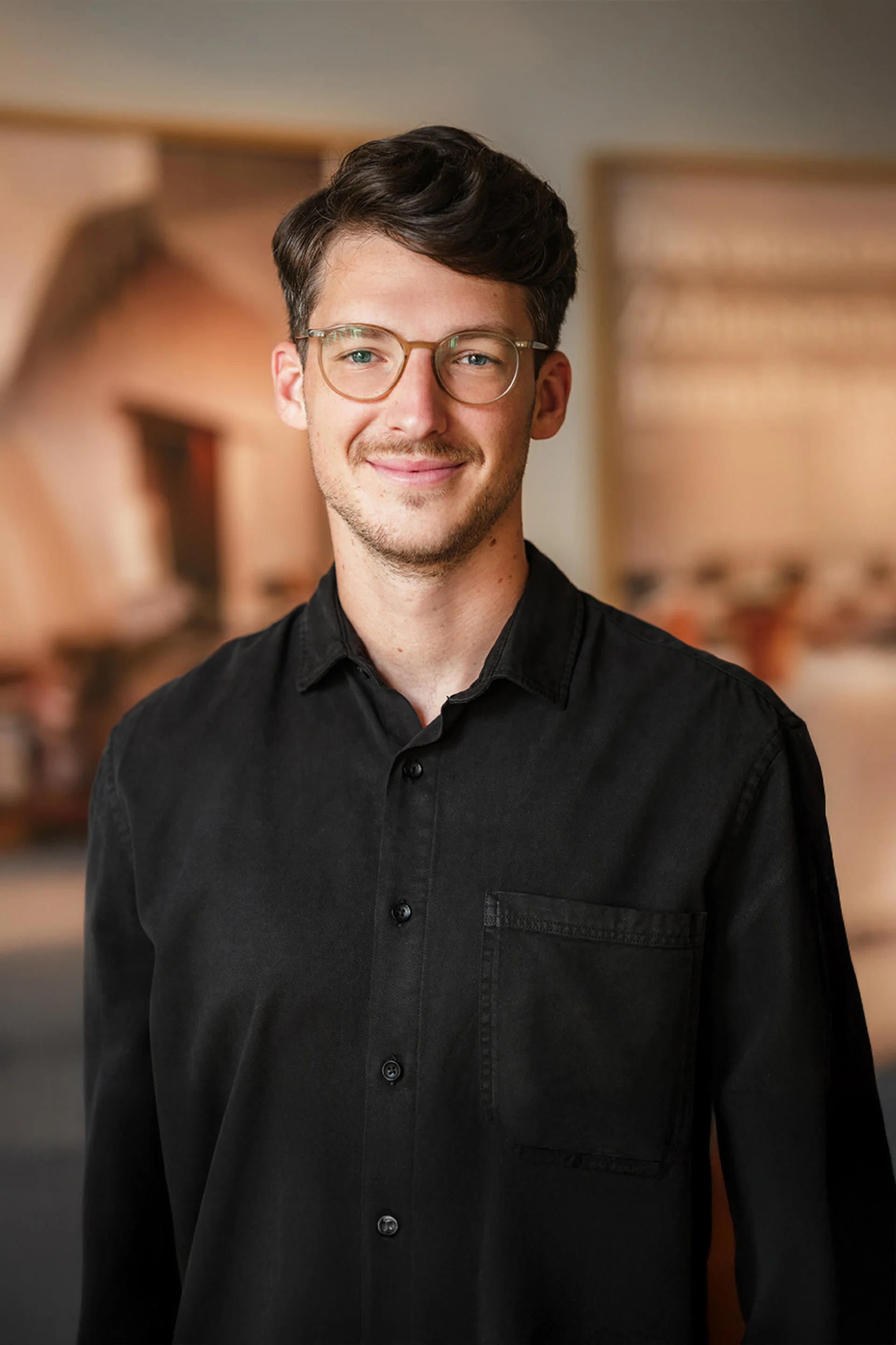 Ein junger Mann mit Brille und schwarzem Hemd steht in einem Innenraum und lächelt in die Kamera.