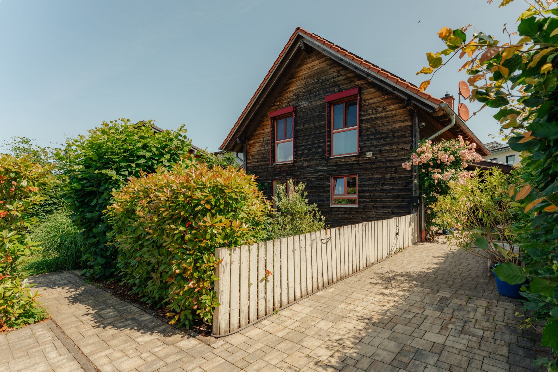 Holzhaus mit rotem Dach, Garten, Zaun und Pflasterweg, umgeben von grünen Büschen.