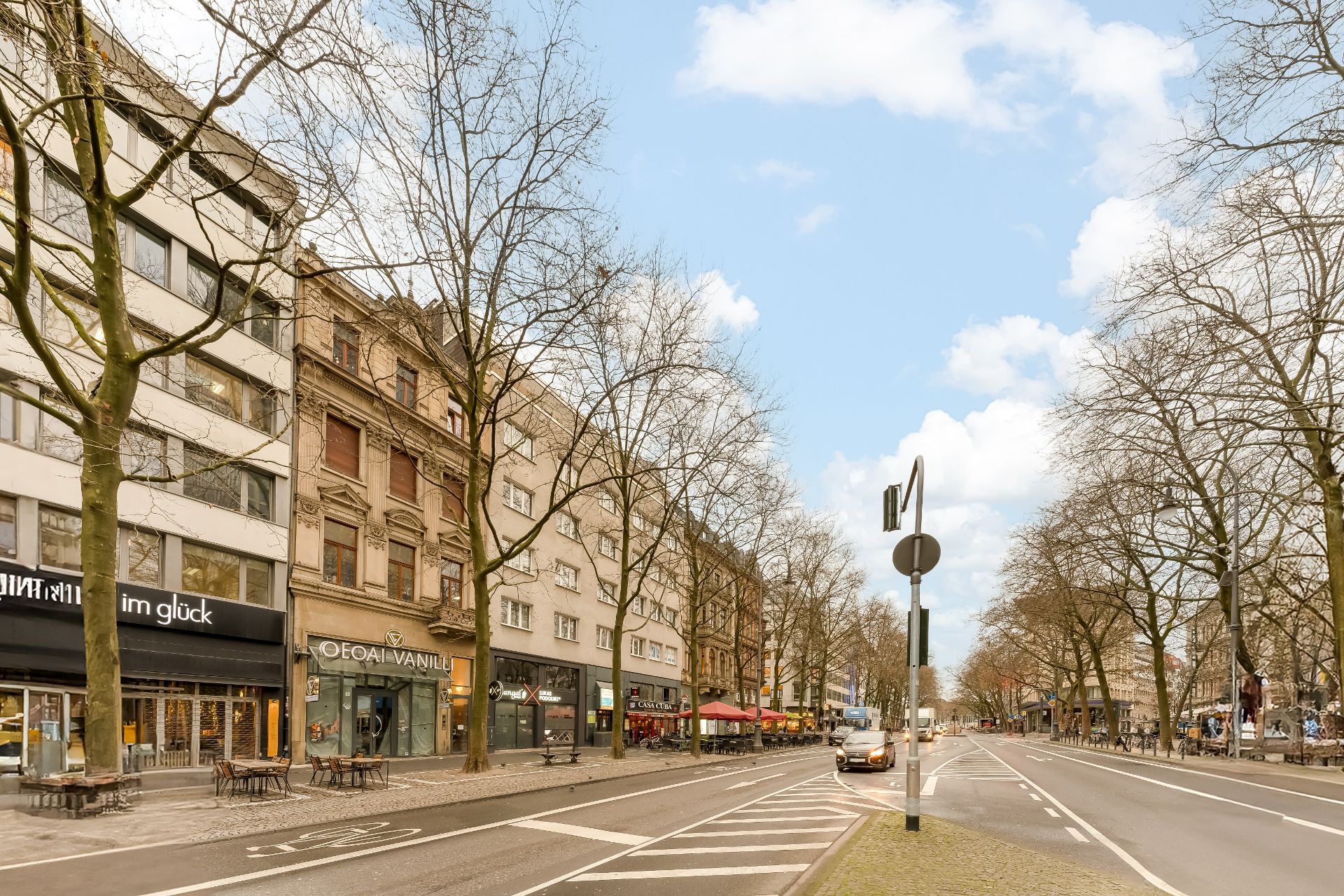 Straßenszene mit Wohnhäusern, Geschäften, Bäumen und Autos an einer breiten Straße.